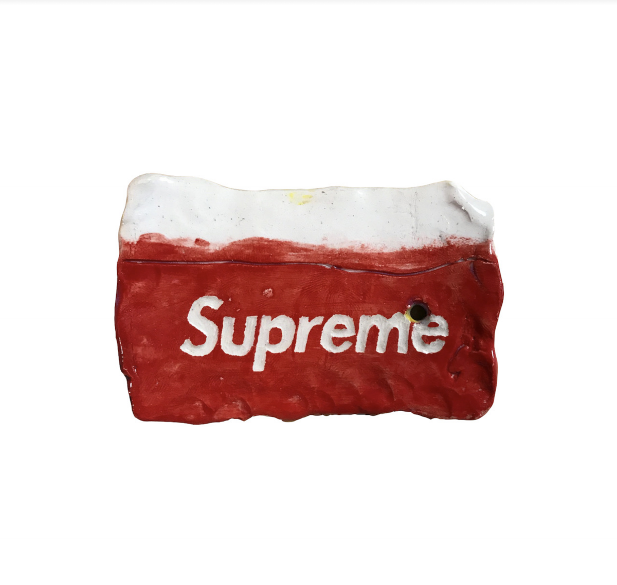 A red and white ceramic object resembling a worn metro card, with the word "Supreme" printed in white across its red surface. The piece is crafted from clay with glazed finish, showing deliberate signs of distress and weathering, with a small hole punctured through one side.