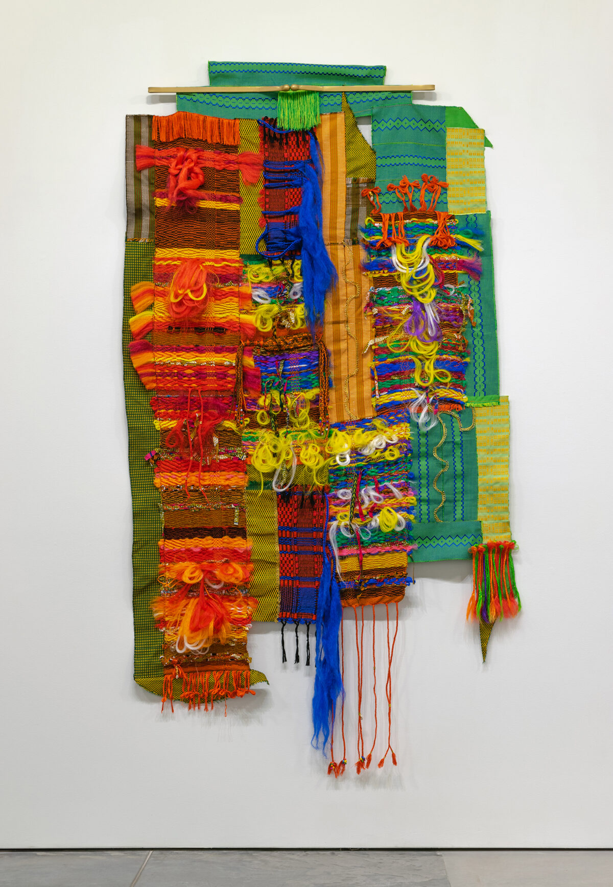 Vibrant textile wall hanging composed of layered fabric strips in shades of red, orange, yellow, green, and blue, incorporating woven patterns, fringe elements, and dangling decorative fibers. The asymmetrical composition features contrasting textures and materials arranged vertically against a white gallery wall.