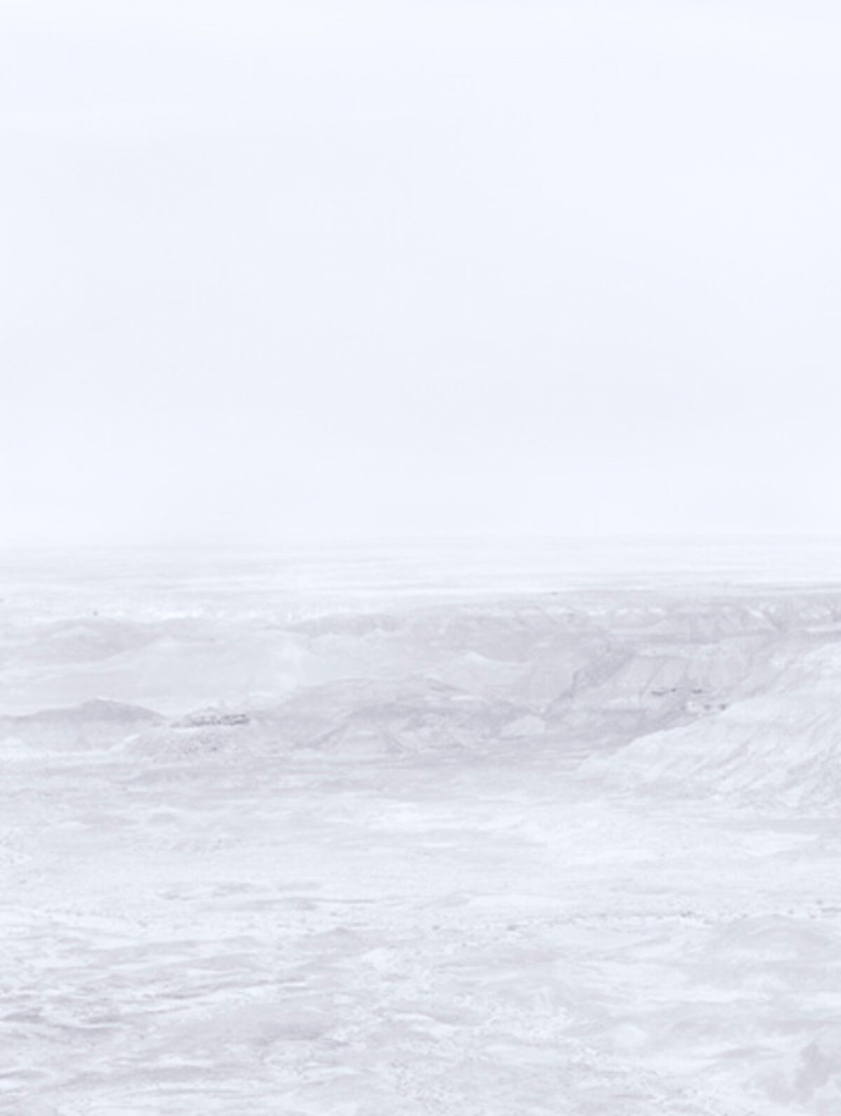 Minimalist landscape featuring layered, undulating terrain in soft whites and grays beneath a pale, overcast sky. The composition emphasizes subtle tonal variations and atmospheric haze across an expansive, largely featureless vista.