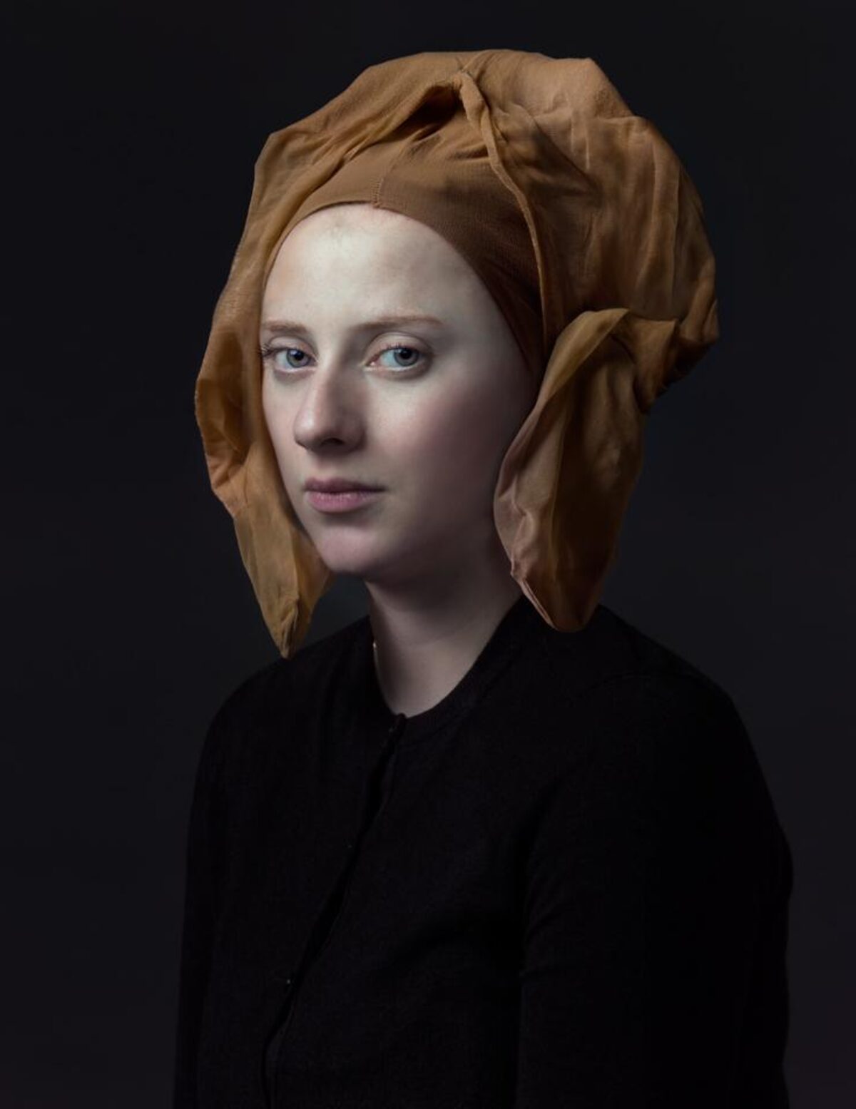 Portrait of a pale-skinned figure with a shaved head wearing a tan linen headwrap and black garment, photographed against a dark background with subtle lighting that emphasizes the sculptural quality of the face and the textural folds of the fabric.