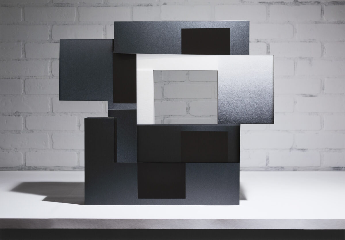 Layered rectangular panels in black, dark gray, and white arranged in an overlapping, three-dimensional composition against a white brick wall. The stacked forms create depth through varying planes and tones, with some surfaces appearing textured or printed, casting subtle shadows on the white surface beneath.
