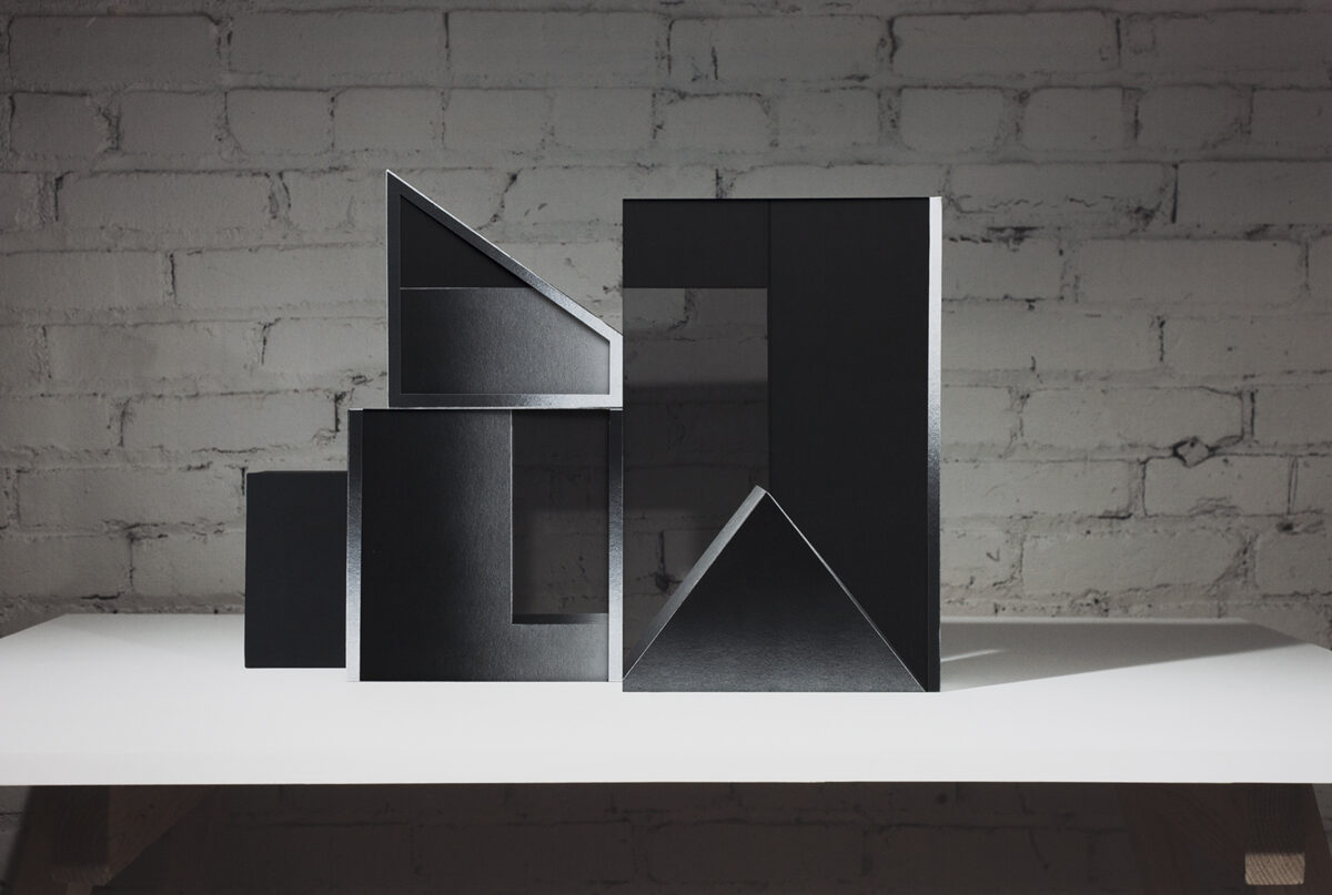 Abstract geometric sculpture made of black layered materials arranged in ascending rectangular and triangular forms on a white platform. The composition features sharp angles, cutouts, and overlapping planes that create dramatic shadows against a weathered brick wall.