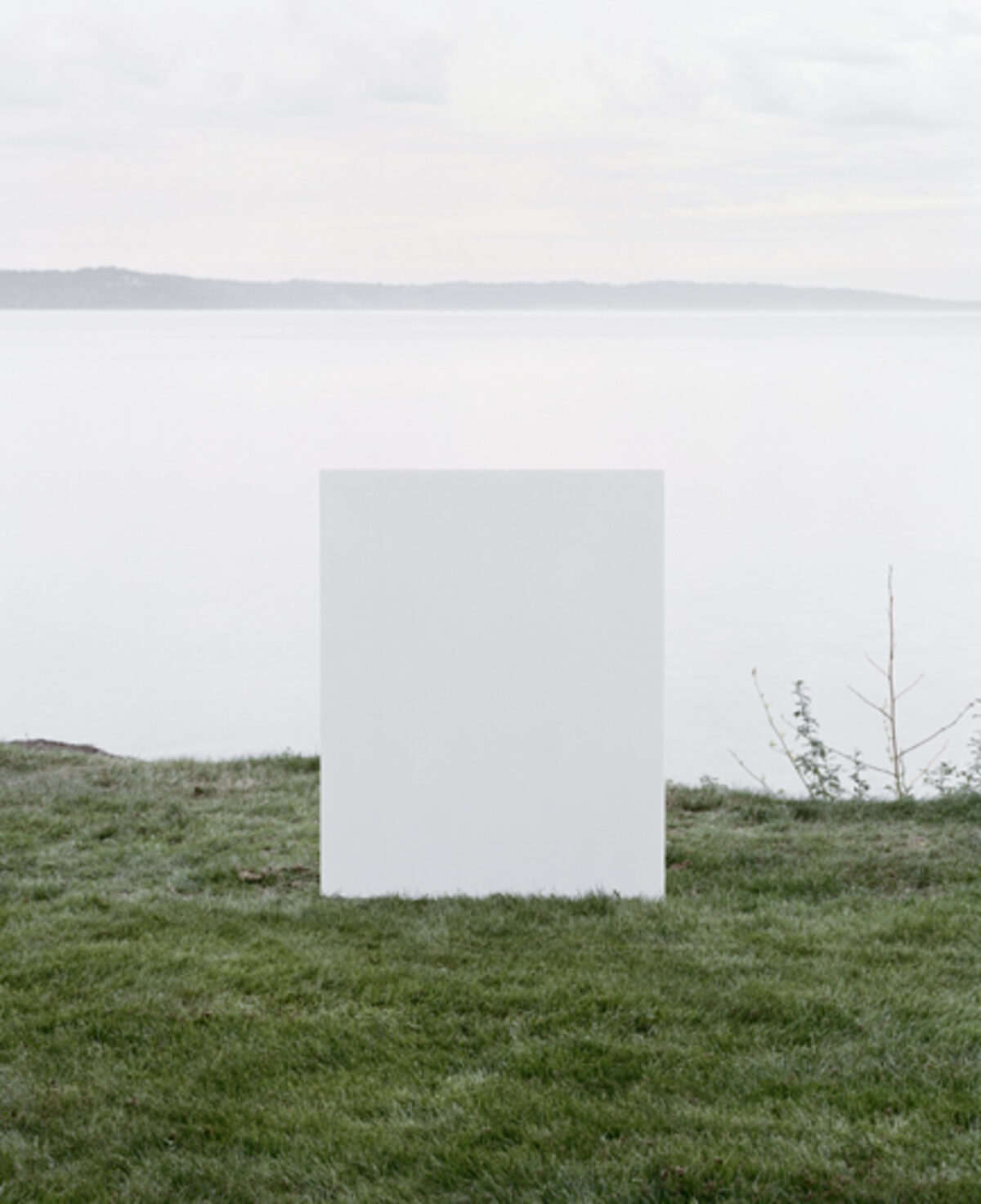 A large white rectangular form stands centered in a grassy field overlooking a calm body of water and distant shoreline under an overcast sky. Sparse dried vegetation and a leafless branch appear to the right of the geometric structure.