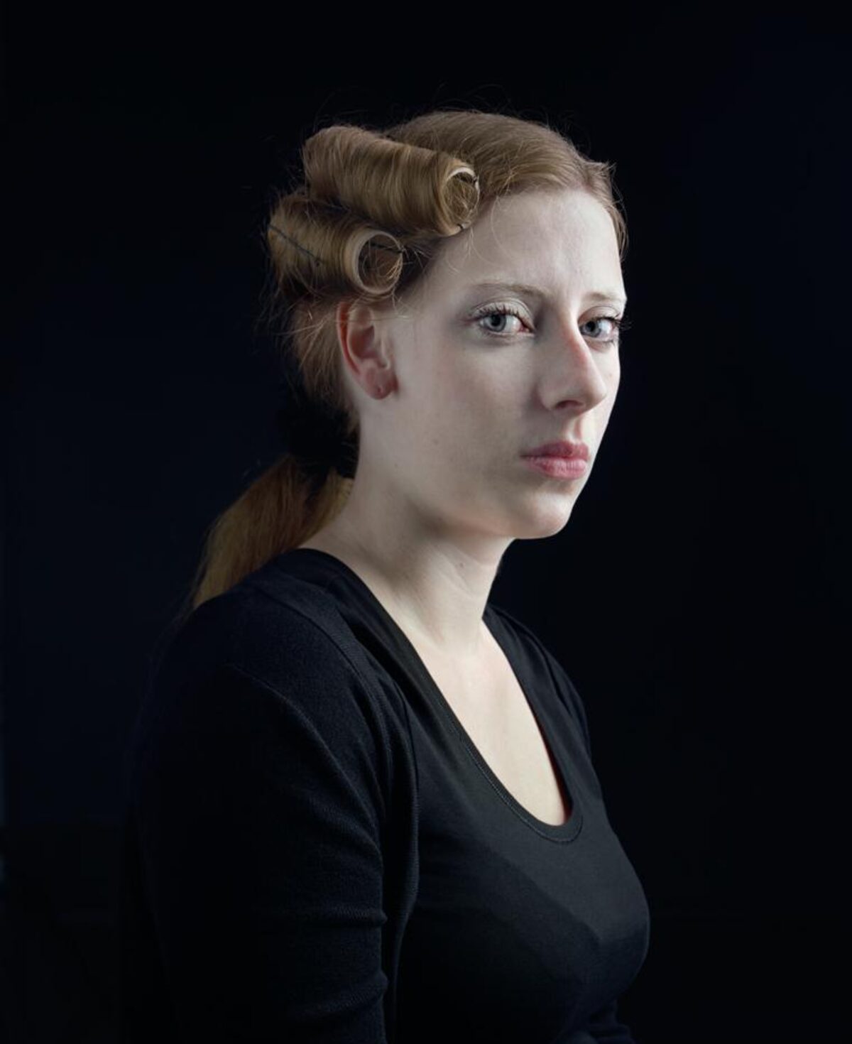 Portrait of a woman in profile against a black background, wearing a black long-sleeved top. Her hair is styled in vintage waves and pinned up on one side, with a ponytail visible. Her gaze is directed downward and to the side, creating a contemplative mood. The lighting is soft and controlled, emphasizing the delicate features and pale complexion of her face.