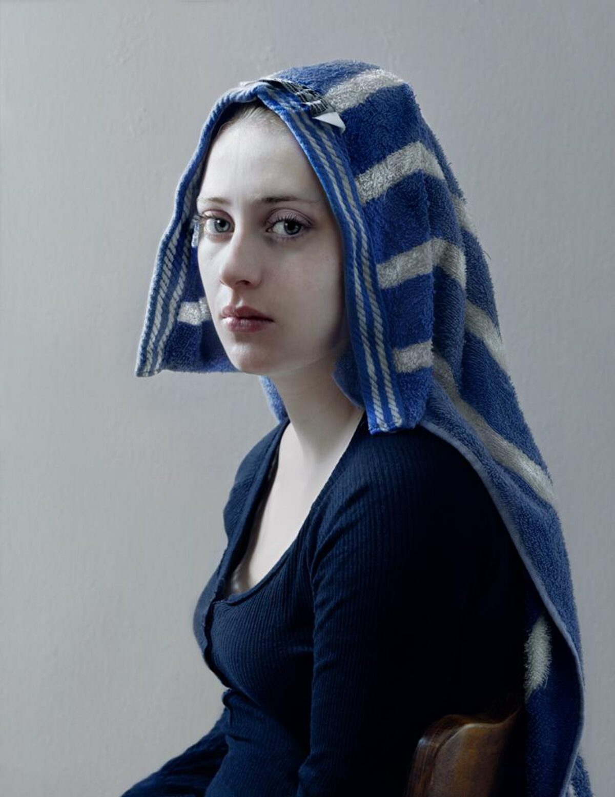 A young woman in profile facing left, wearing a dark ribbed navy garment with a blue and gray striped towel wrapped around her head. She has pale skin, defined facial features, and a composed expression, set against a neutral gray background with warm brown tones visible at the lower right edge.