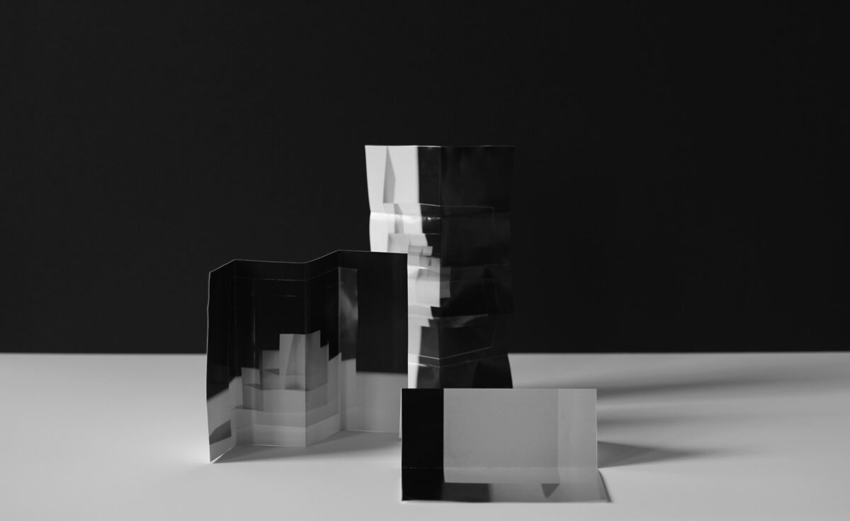 Three-dimensional paper or card structures with black and white surfaces arranged on a light gray floor against a black wall, creating geometric forms with layered planes and sharp angles that cast distinct shadows.