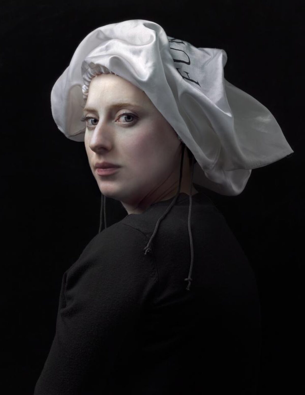 Portrait of a figure shown in three-quarter view against a black background, wearing a black garment with a distinctive white linen headwrap with voluminous folds. The figure gazes toward the viewer with a composed expression, rendered in a highly detailed, photorealistic style with naturalistic skin tones and subtle modeling of light and shadow.