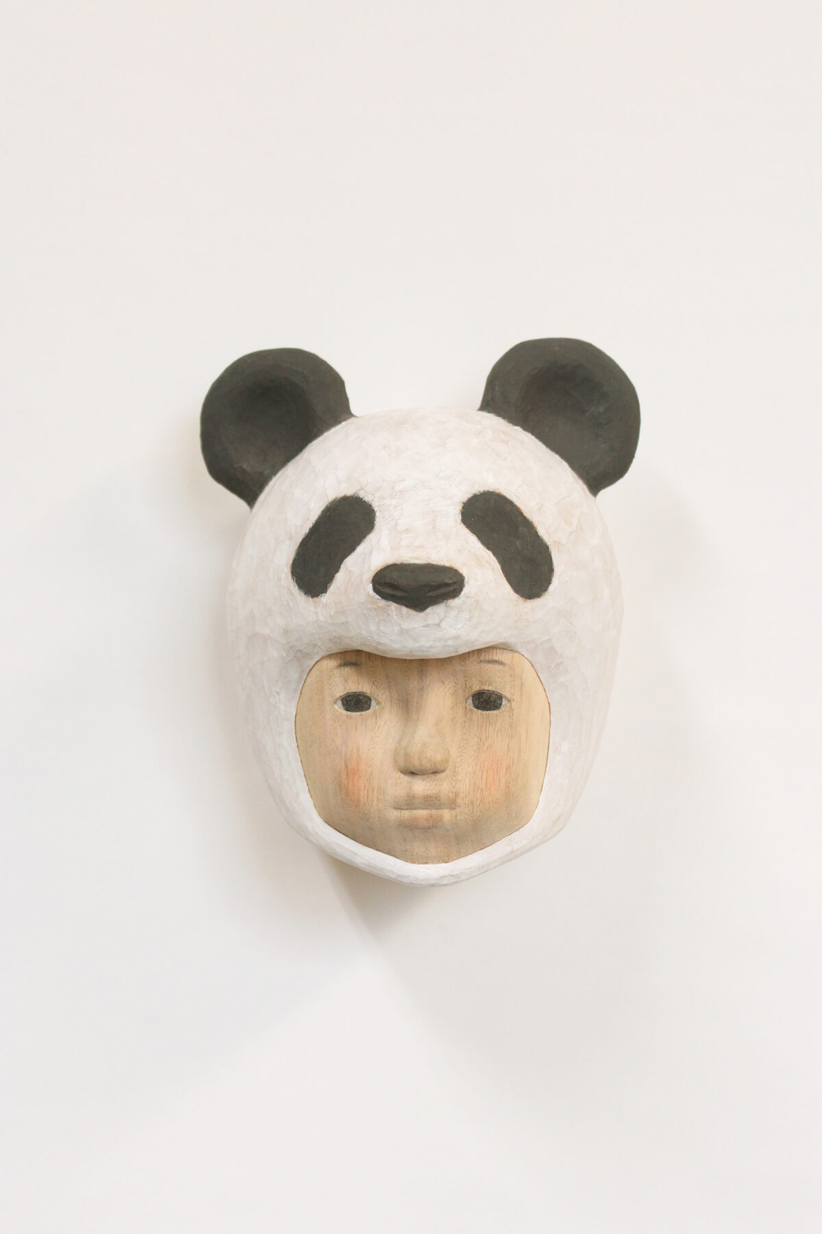 A carved wooden mask depicting a human face wearing a giant panda head covering, with black and white coloring and subtle flesh-toned facial features below. The sculpture combines naturalistic human portraiture with stylized animal characteristics, mounted on a white background.