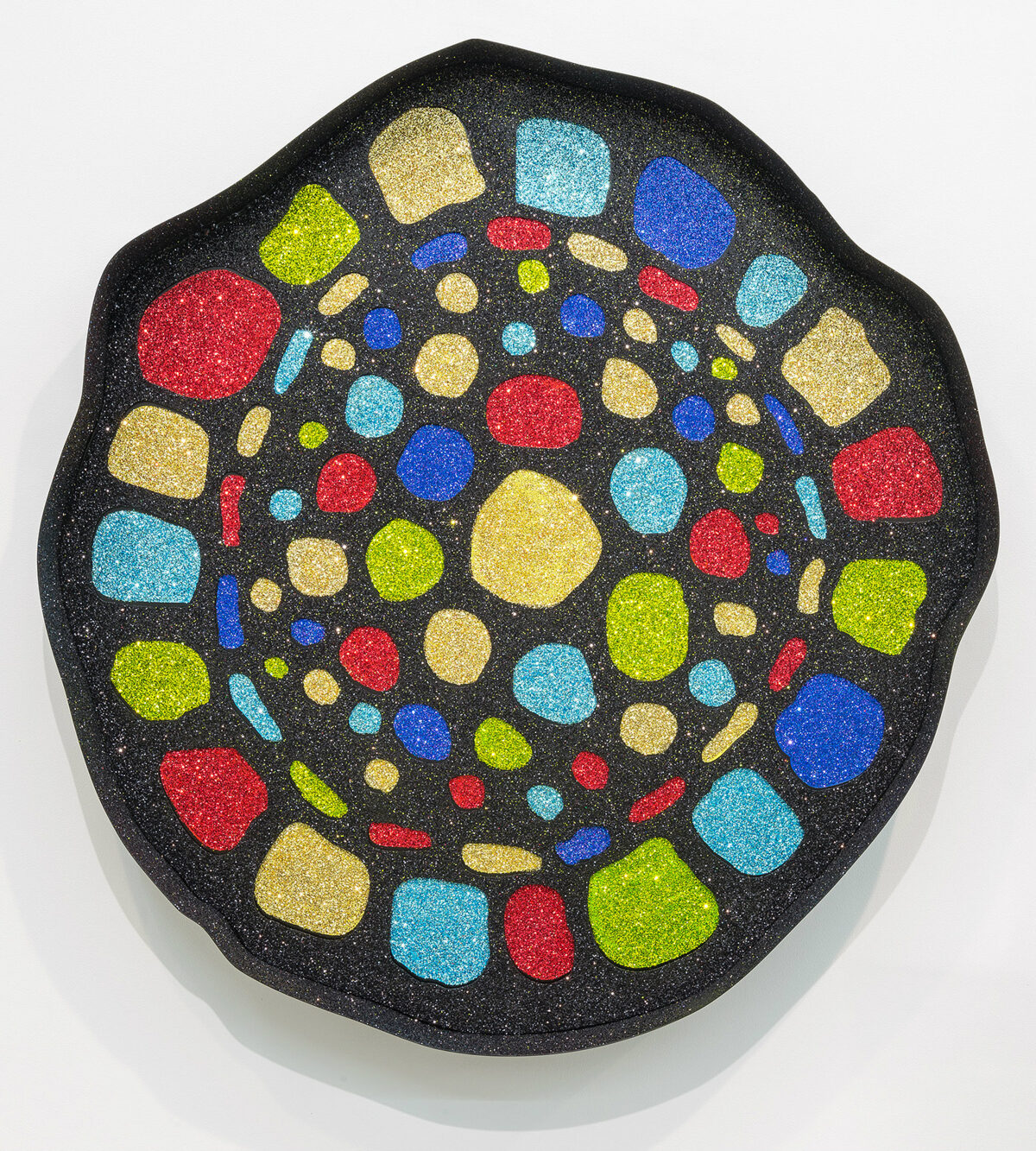 An organic, kidney-shaped black panel densely covered with colorful glittering shapes in red, yellow, blue, cyan, and lime green. The varied forms—ranging from small circles and rectangles to larger rounded and squared shapes—are scattered across the surface with a shimmering, jewel-like finish that creates an energetic, mosaic-like composition.