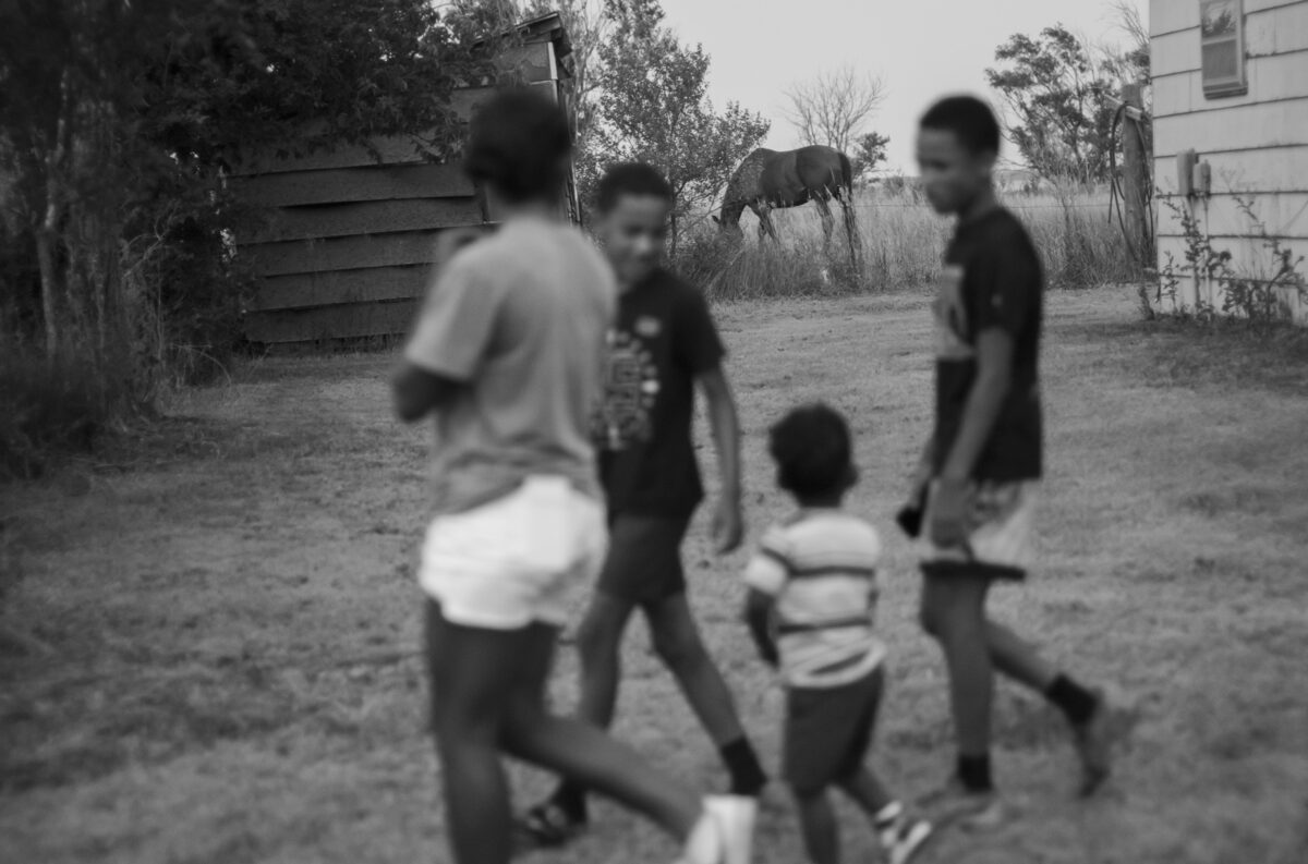 Black and white photograph of several figures in motion in a rural dirt yard between weathered wooden structures and overgrown vegetation. The subjects are intentionally blurred, creating a sense of movement and ethereal quality, while the architectural elements and landscape remain in sharper focus.