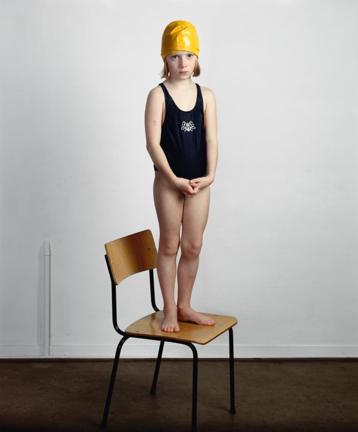A young child in a black swimsuit and bright yellow swimming cap stands barefoot on a wooden chair with black metal legs, positioned against a white wall. The child's hands are clasped in front, gazing directly at the camera in a contemplative pose.