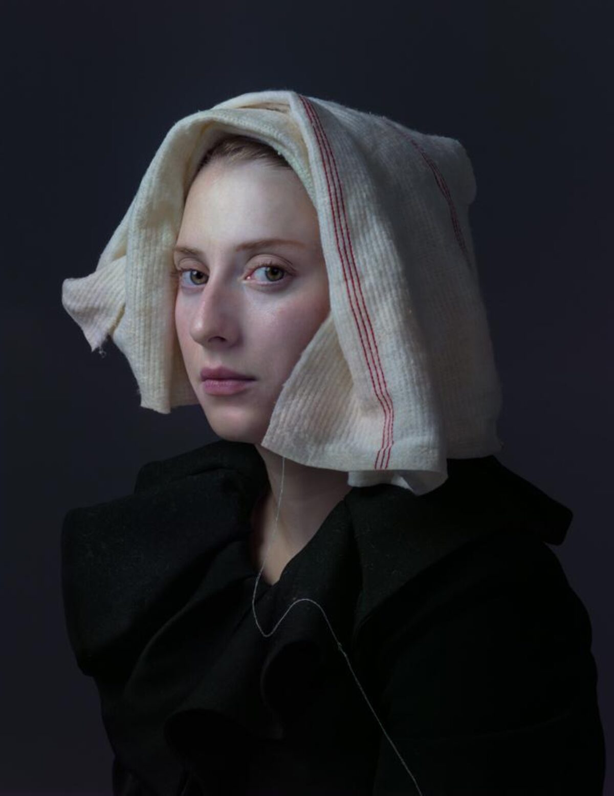 Portrait of a young woman against a dark background, wearing a black garment and a cream-colored linen head covering with a red stripe. Her gaze is directed slightly away from the camera, with a composed, contemplative expression.