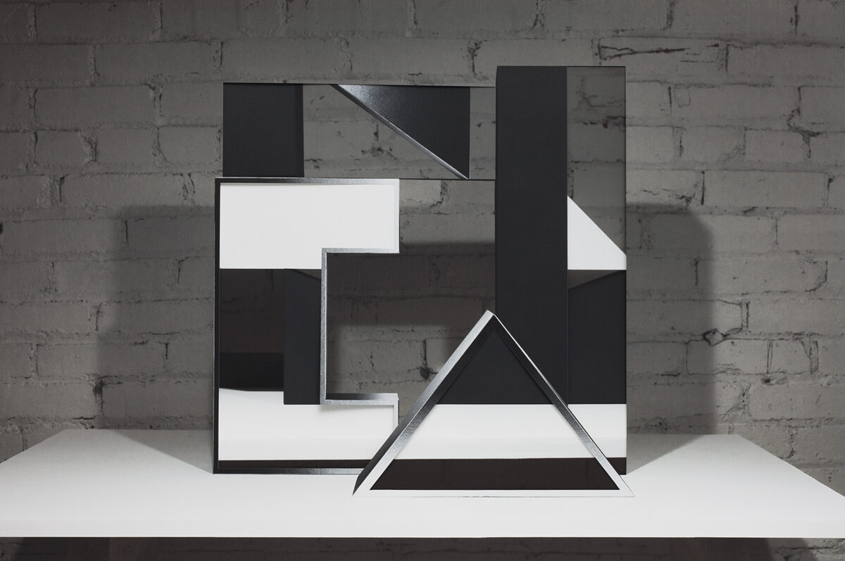 Abstract geometric composition of layered black and white paper forms arranged on a white platform against an exposed brick wall. Interlocking rectangles and a triangular shape create a three-dimensional collage with sharp angles and negative space, emphasizing contrast between dark and light surfaces.