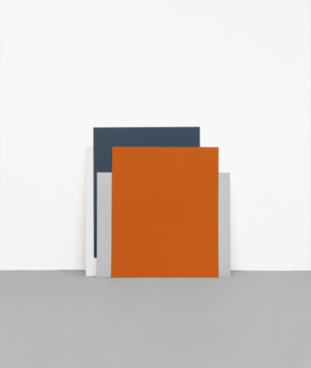 Abstract geometric composition featuring overlapping rectangular forms in dark gray, orange, and light gray tones arranged against a gray ground, creating a minimalist architectural or sculptural arrangement.