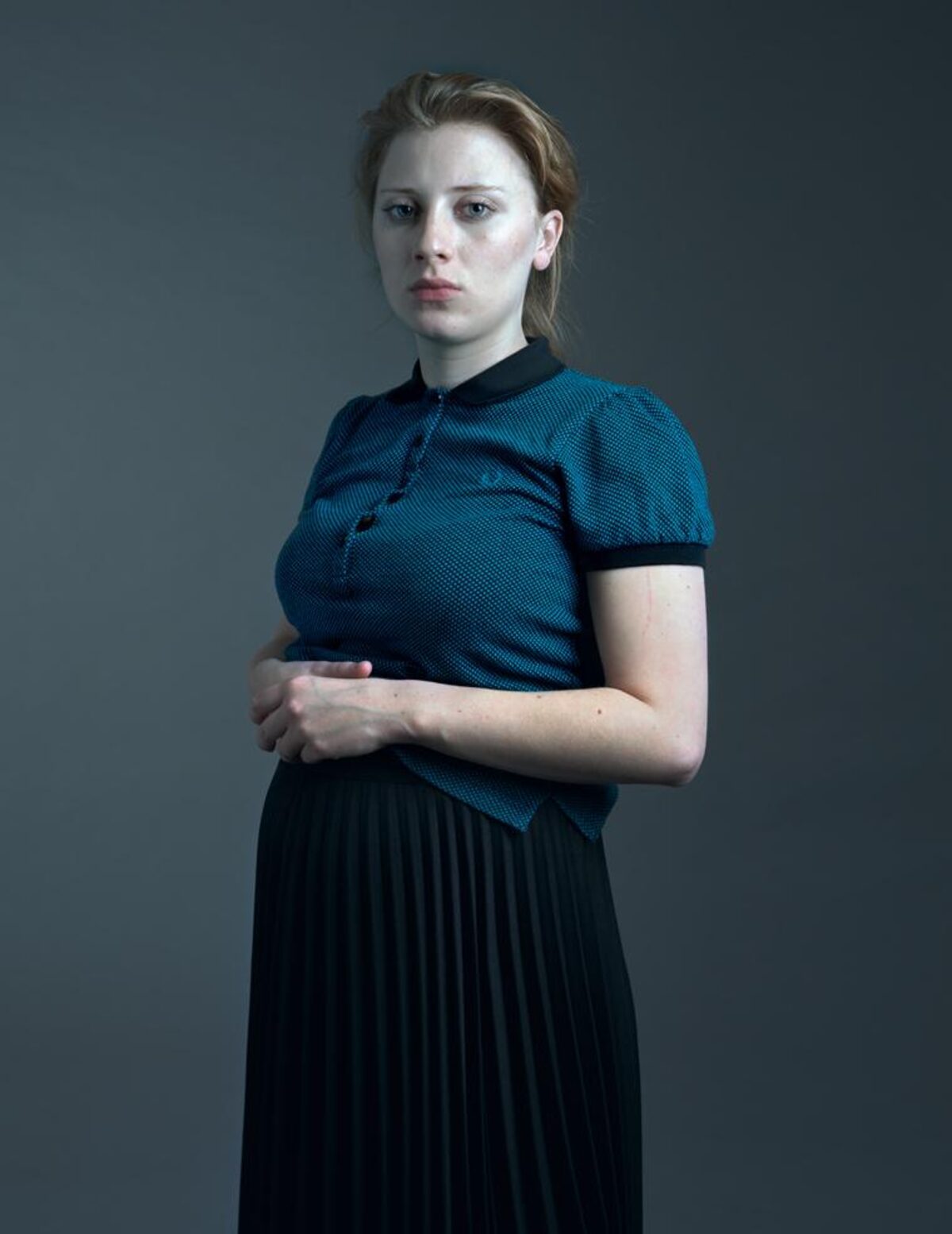A woman with reddish-blonde hair stands against a neutral gray background, wearing a teal textured short-sleeved top with a black collar and a black pleated skirt. She gazes directly at the camera with a composed, contemplative expression, her arms crossed in front of her body.