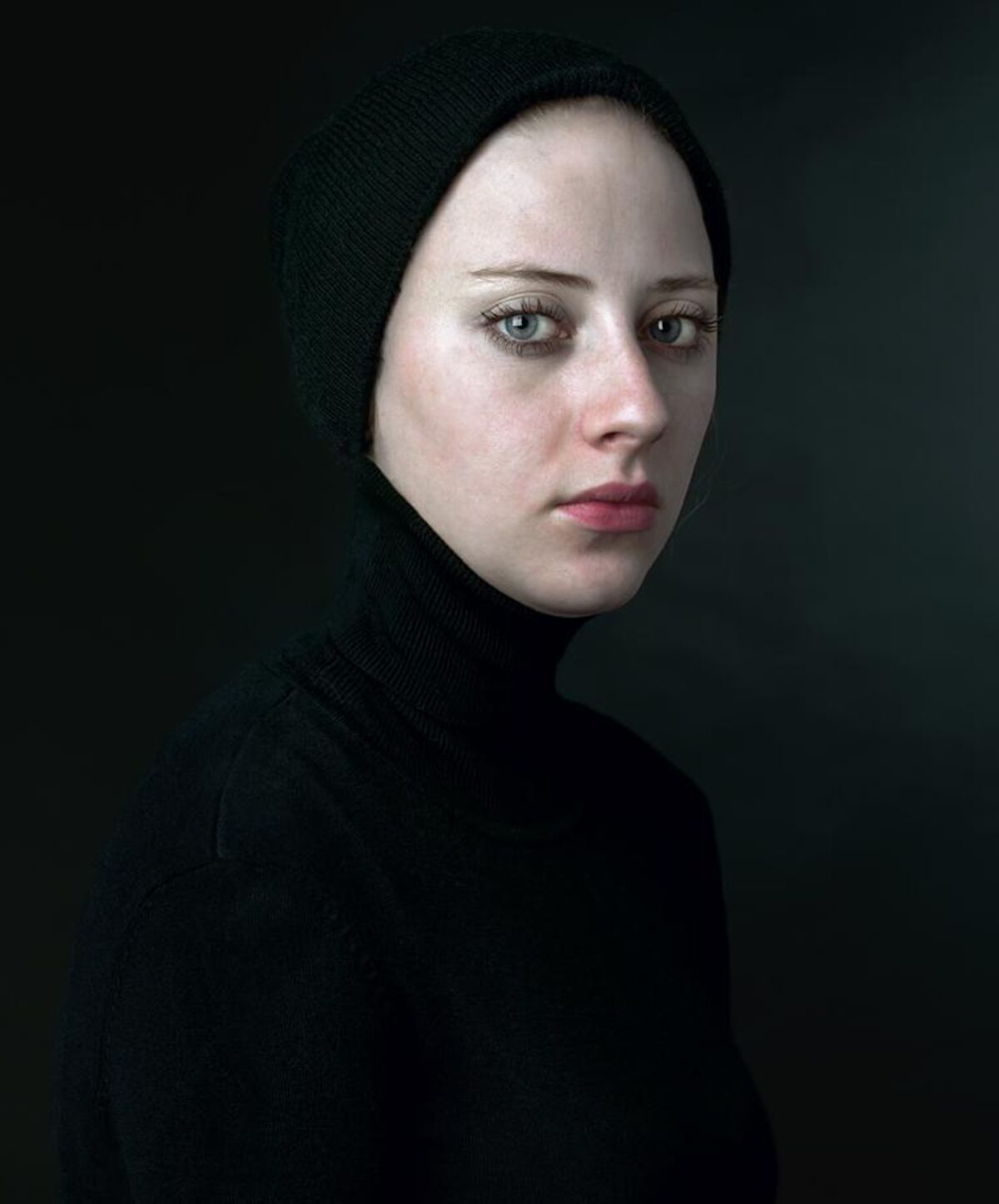 Portrait of a pale-skinned person wearing a black knit hood and turtleneck, photographed against a dark background in three-quarter view. The subject gazes downward with a contemplative expression, their face luminously lit against the shadowed composition.
