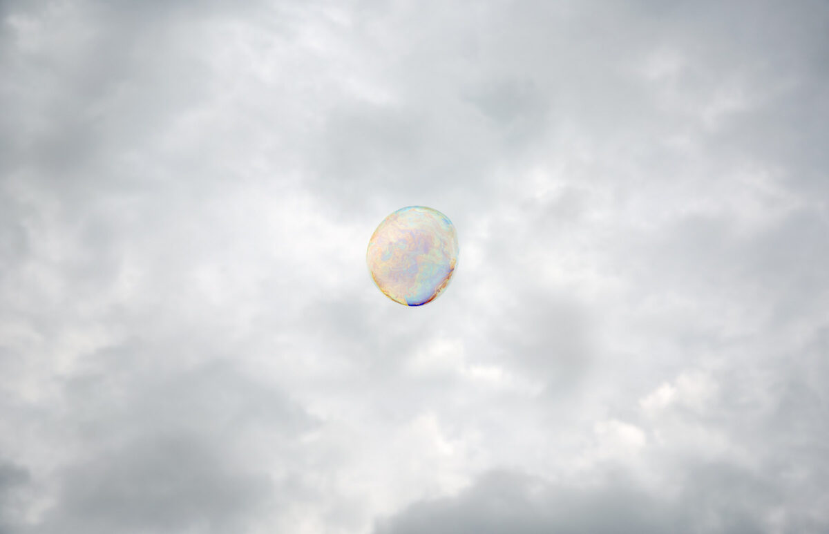 A translucent iridescent sphere floats against a soft, overcast sky filled with white and gray clouds. The bubble's delicate surface displays subtle shifts of pastel color—pale blues, pinks, and yellows—characteristic of light refracting through a thin film.