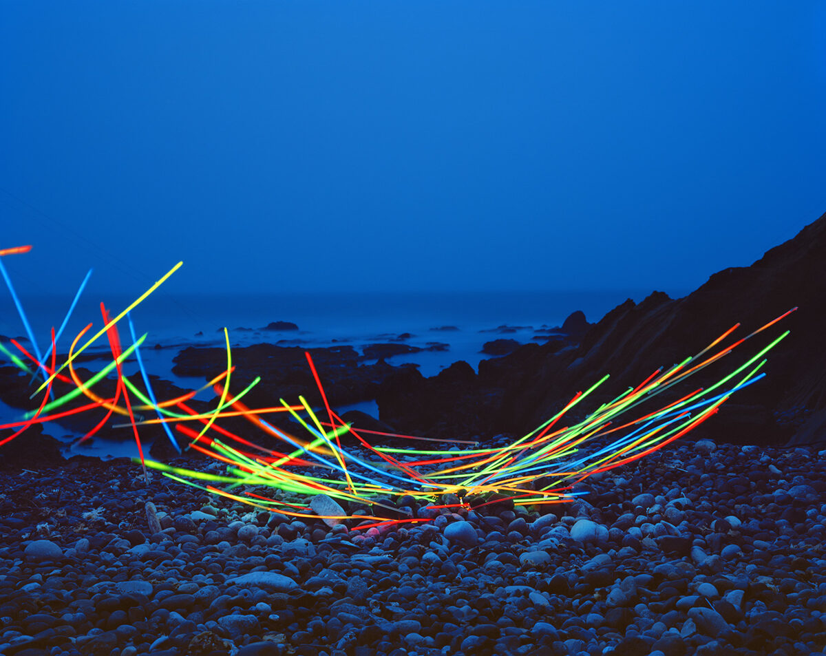 Neon glow sticks arranged in tangled, radiating patterns across a rocky beach at dusk, their bright red, yellow, green, and blue lines creating dynamic linear forms against the darkened landscape and twilight sky.