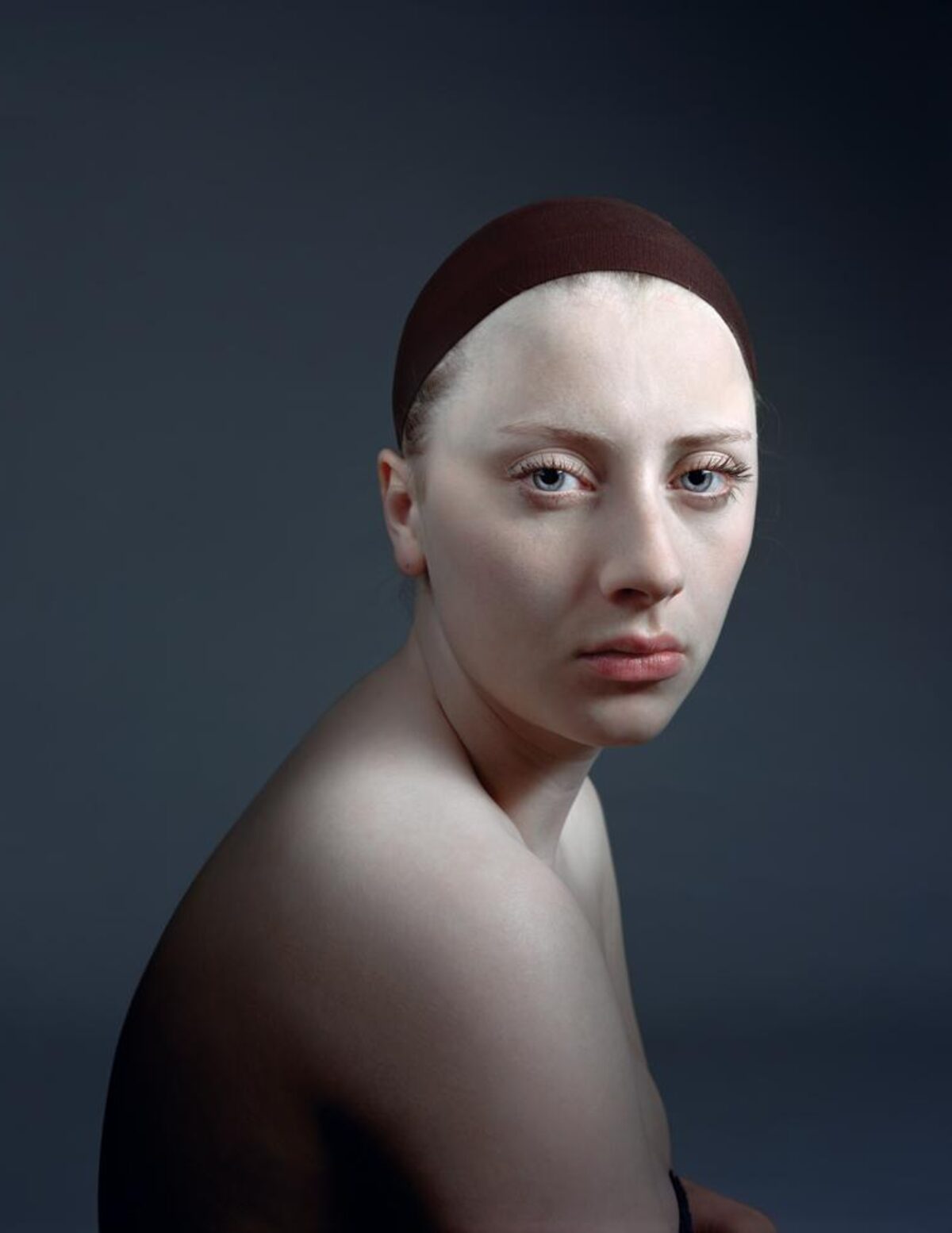 Portrait of a pale-skinned figure with shaved head wearing a dark maroon headband, shown in three-quarter view against a cool blue-gray background. The figure has a contemplative expression and bare shoulders, rendered with precise detail and smooth modeling of form.