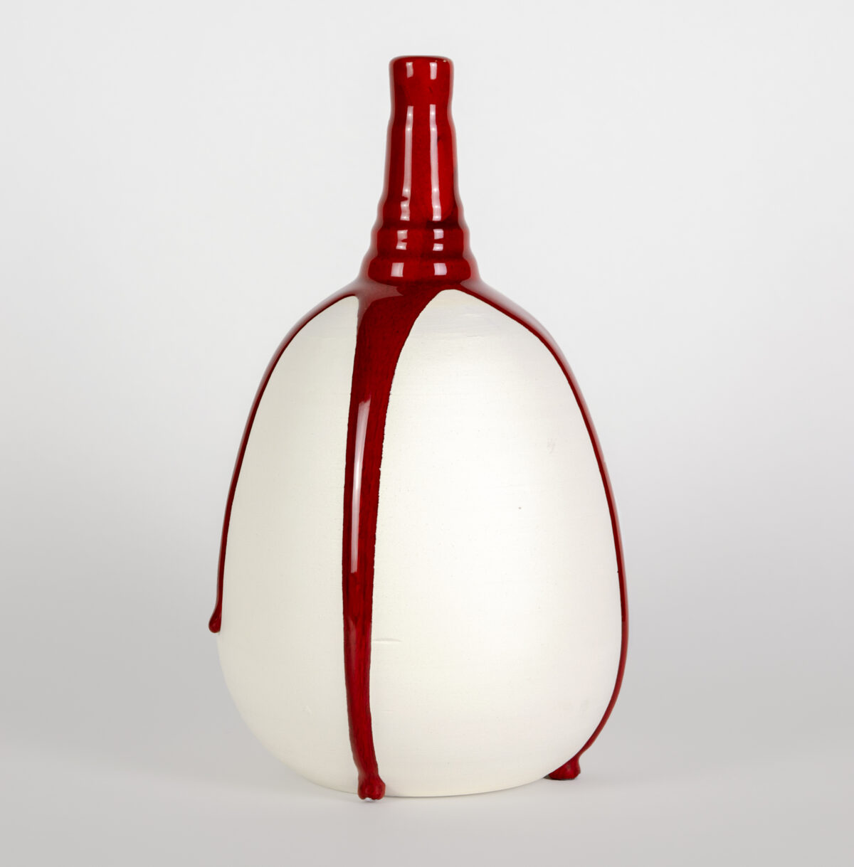 A rounded, cream-colored vessel with a tapered red neck and spout, bound vertically by bold red straps that extend from top to bottom, creating a striking contrast between the soft neutral body and vibrant crimson accents.