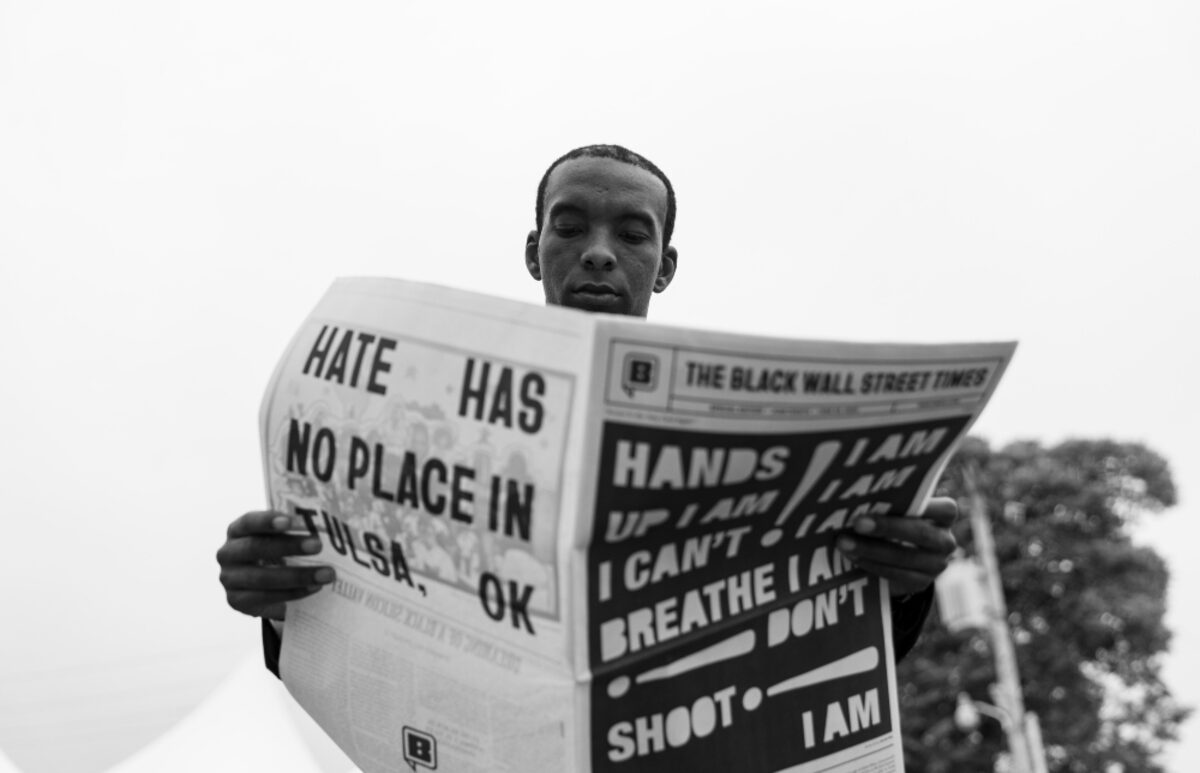 Black and white photograph of a young man holding up a newspaper with protest headlines including "Hate Has No Place in Tulsa, OK" and "I Can't Breathe." The composition is shot from a low angle against a light sky, with trees visible in the background.