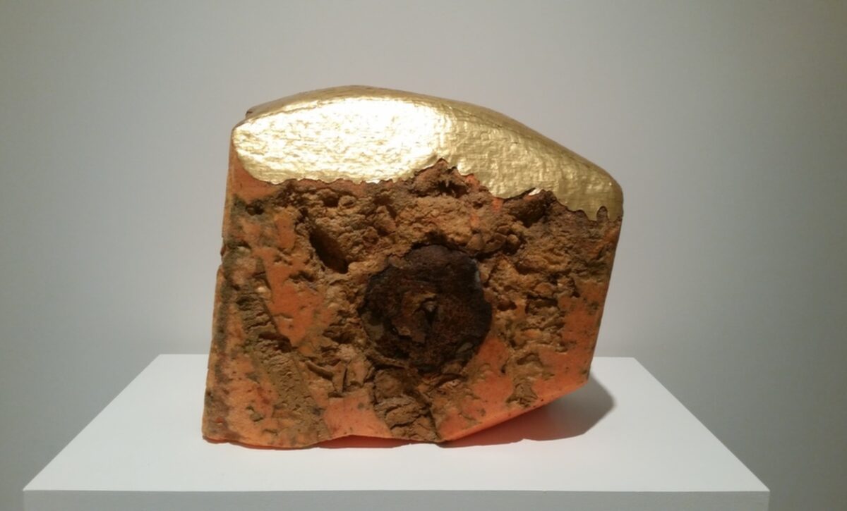 A sculptural form composed of reddish-brown textured material with an irregular, eroded surface sits on a white platform. The top half is covered in burnished gold leaf, creating a stark contrast with the rough, debris-like lower portion.