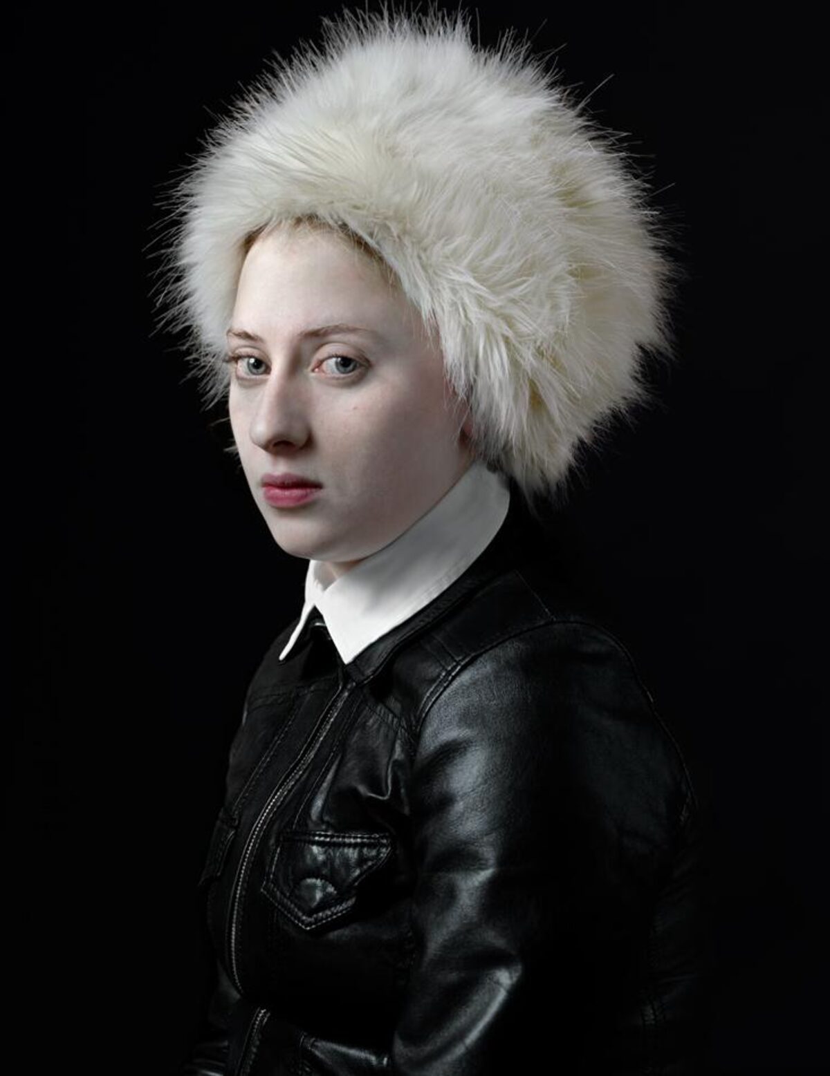 Portrait of a person with pale complexion wearing a voluminous white fur hat and black leather jacket with white collar against a dark background, shown in three-quarter view facing left.