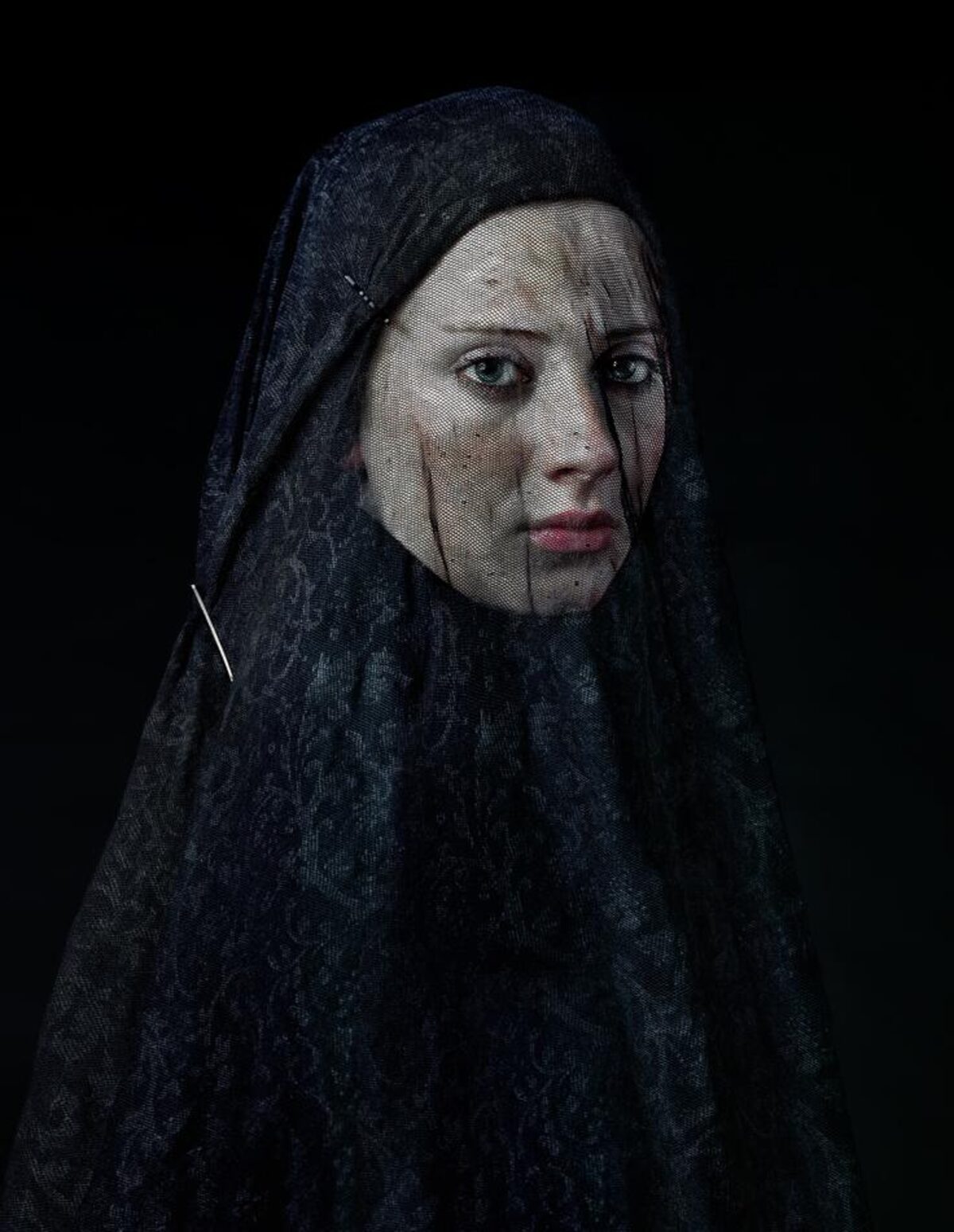 Portrait of a person wearing a dark, textured head covering against a black background. The figure gazes directly toward the camera with a composed expression, their pale face contrasting sharply with the deep navy fabric that drapes over their head and shoulders.