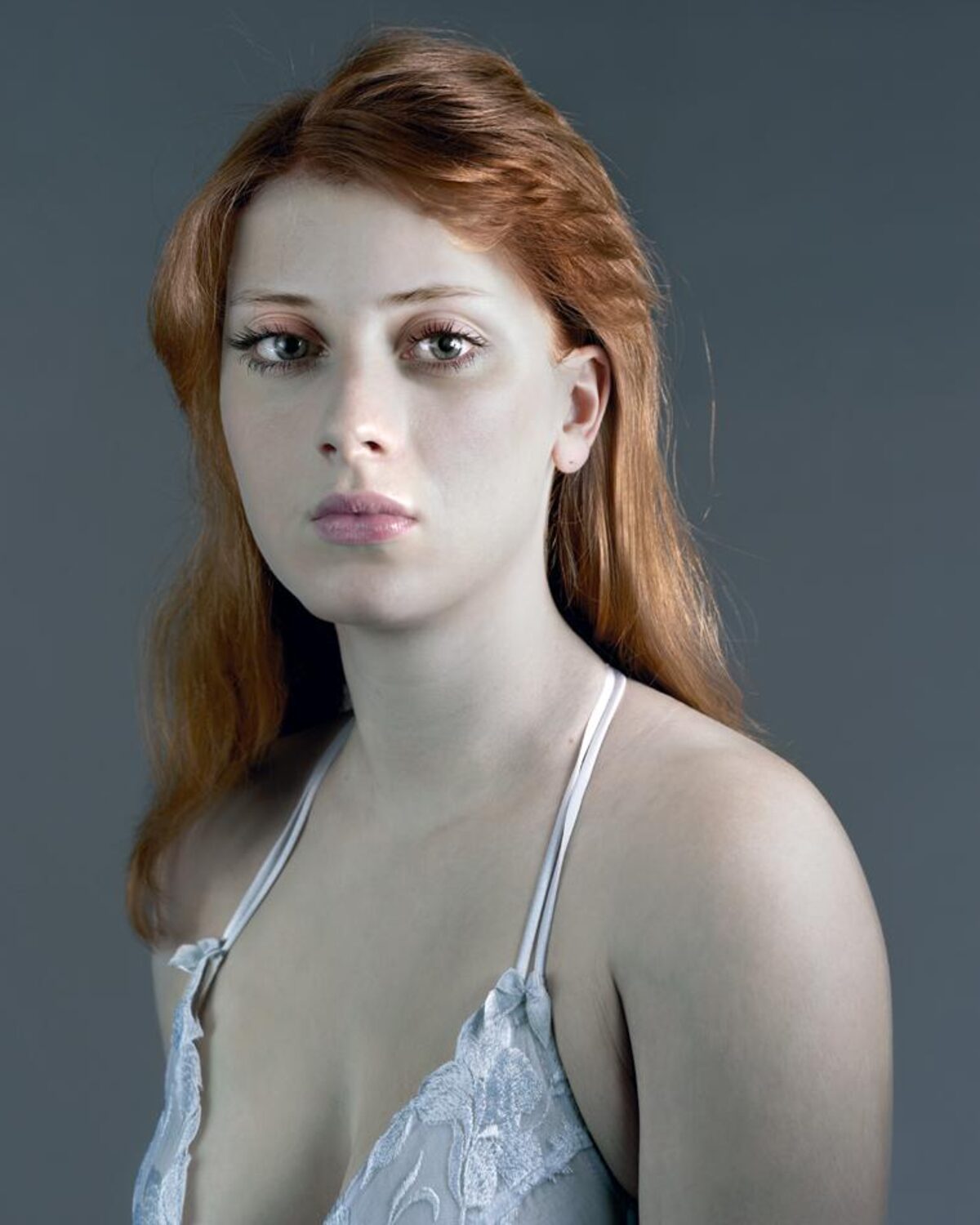 Portrait of a woman with long reddish-brown hair gazing downward, wearing a light blue lace halter top against a neutral gray background. The photograph employs soft, directional lighting that emphasizes the subject's pale complexion and contemplative expression.
