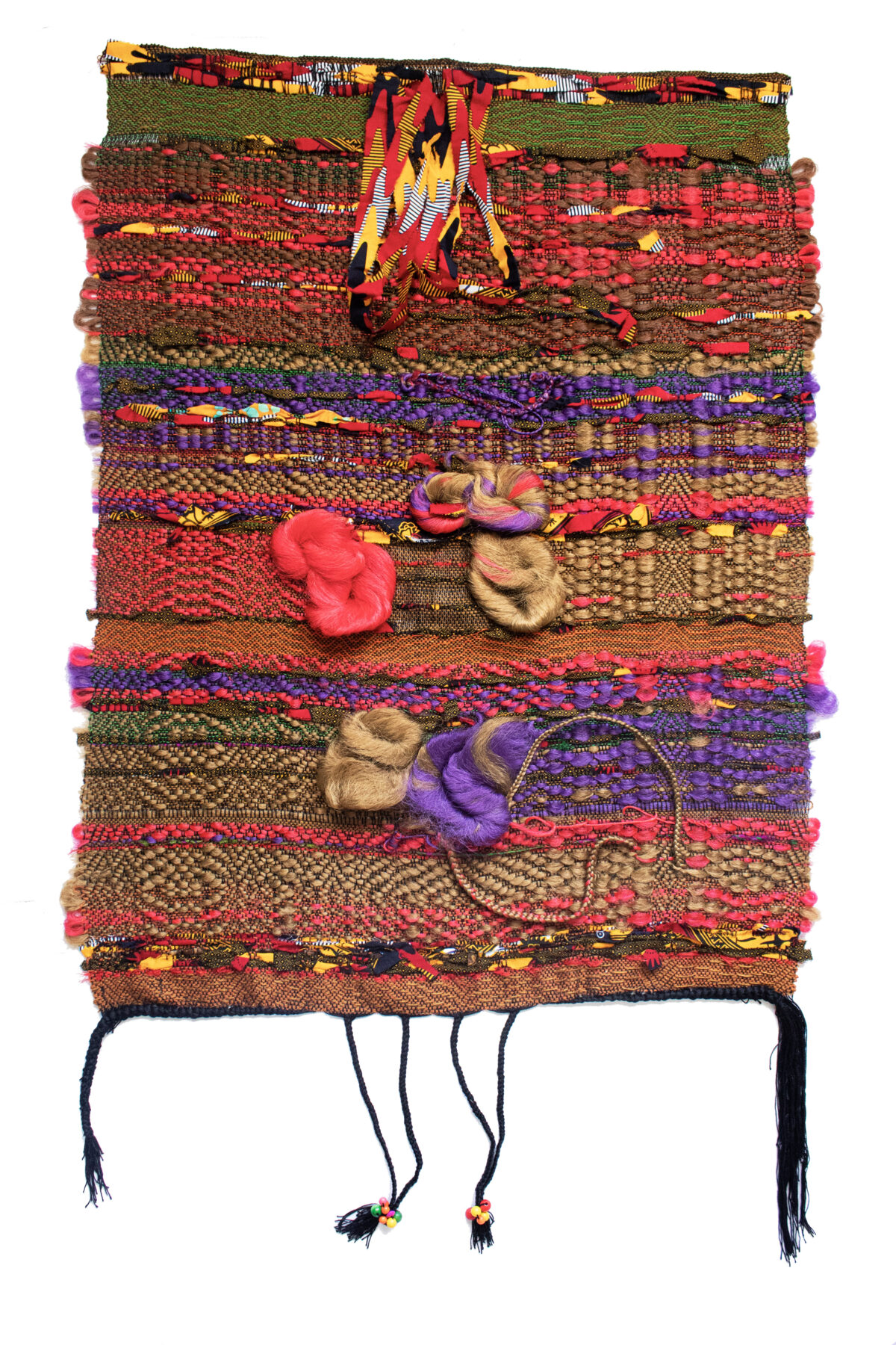 Vertically stacked horizontal bands of richly textured woven fabrics in warm earth tones, vibrant reds, purples, and golds, interspersed with three-dimensional sculptural forms in coral, tan, and purple. Two dark braided cords with beaded ends hang from the bottom of the composition. The layered arrangement combines various textile patterns and materials to create a dense, tactile mixed-media work.