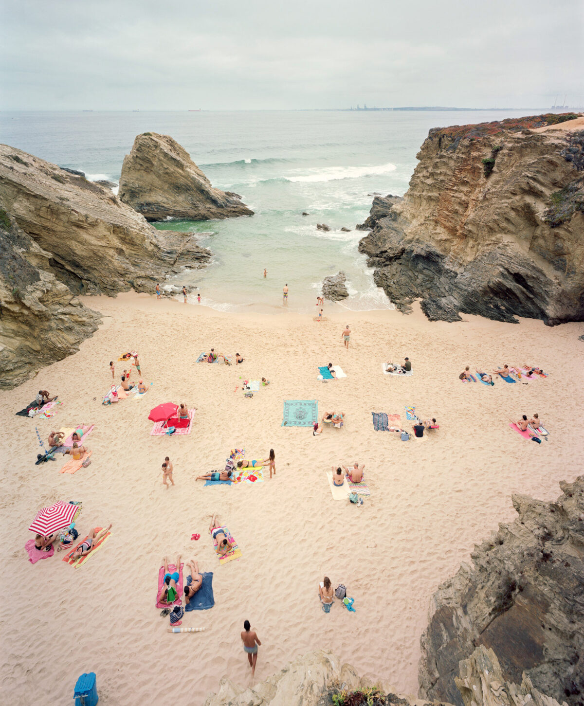 Aerial view of a sandy beach cove enclosed by dramatic rocky cliffs, with scattered beachgoers, colorful umbrellas, and towels dotting the sand. The turquoise and white waters beyond are studded with rocks, under an overcast sky.