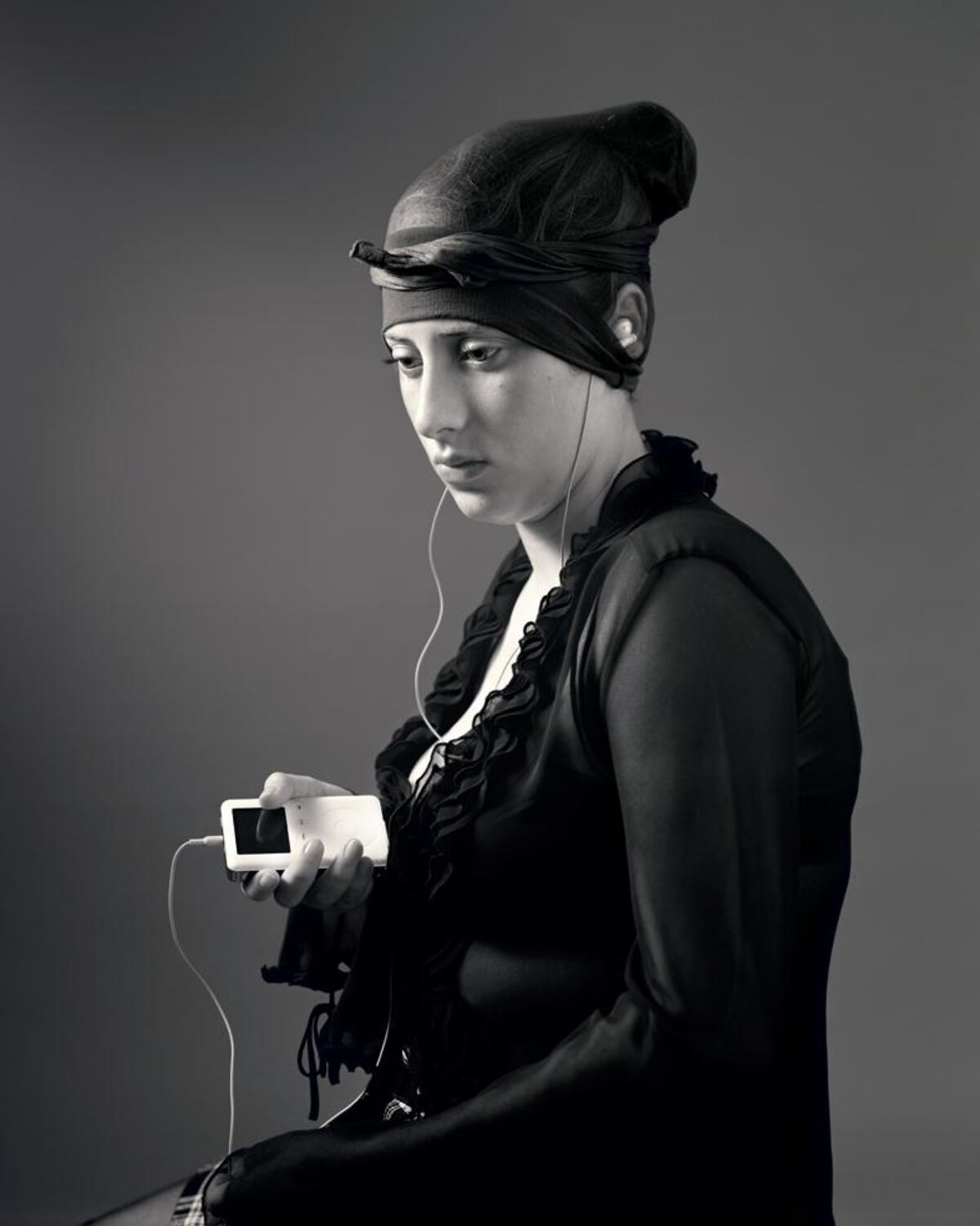 Black and white portrait of a woman in profile wearing a black hooded jacket and headband, holding a small white digital device with white earbuds. The minimalist composition is set against a neutral gray background with dramatic lighting that emphasizes the subject's contemplative expression.