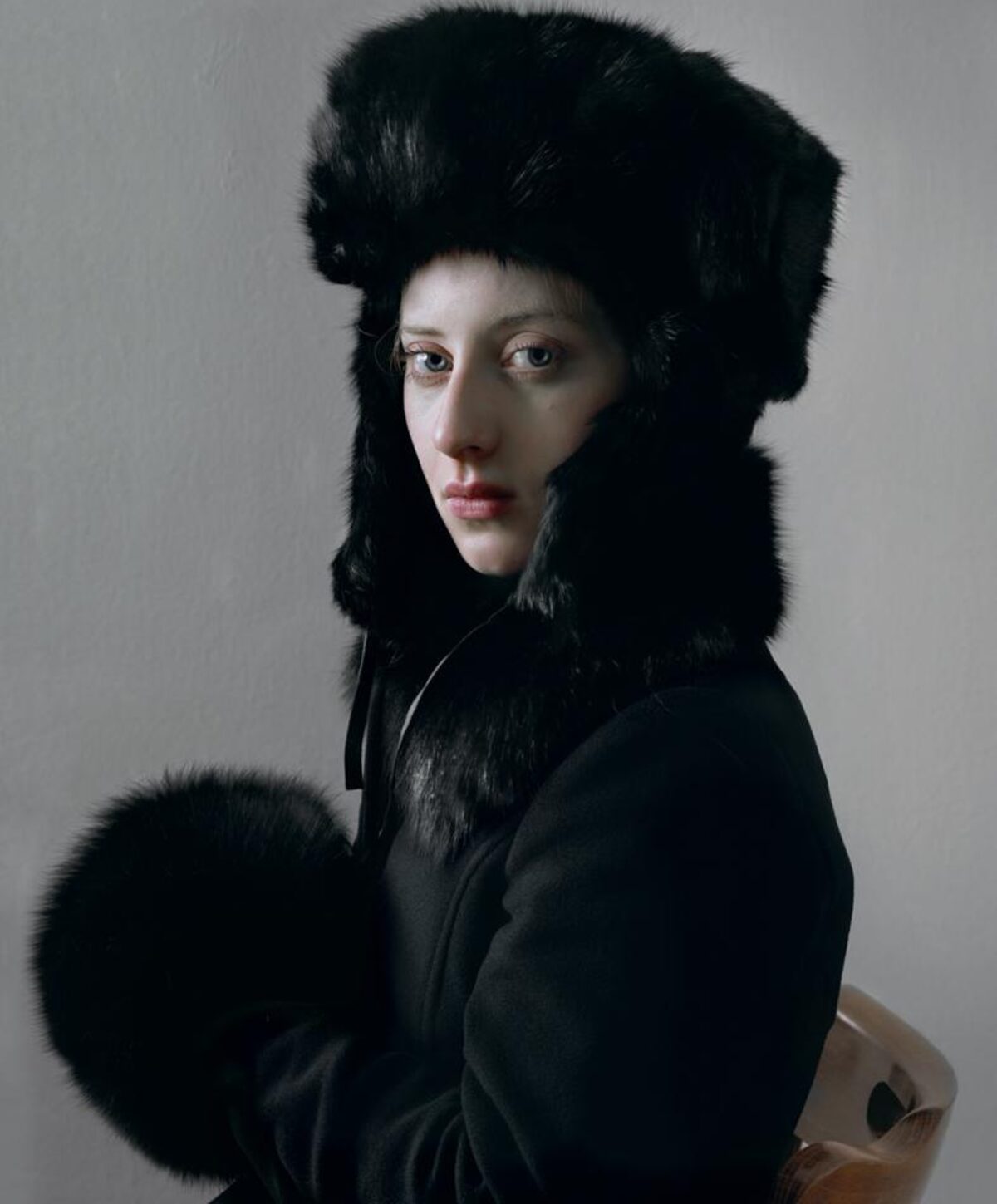 A person with pale complexion gazes over their shoulder while wearing a large black fur hat and collar against a neutral gray background. The subject is dressed in dark clothing with a striking fur muff held at their side, creating a composed portrait with dramatic contrast between the figure's fair skin and the rich black textures of the fur.