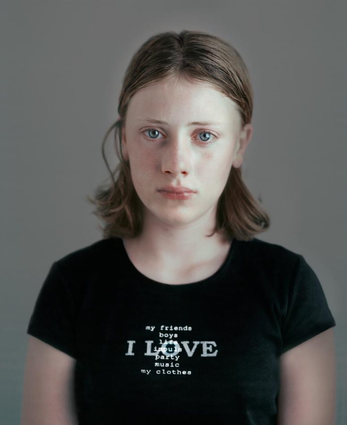 Portrait of a young person with shoulder-length brown hair wearing a black t-shirt printed with white text listing "my friends boys I LOVE party music my clothes" against a neutral gray background. The photograph employs a direct, frontal composition with a contemplative expression and cool color palette.