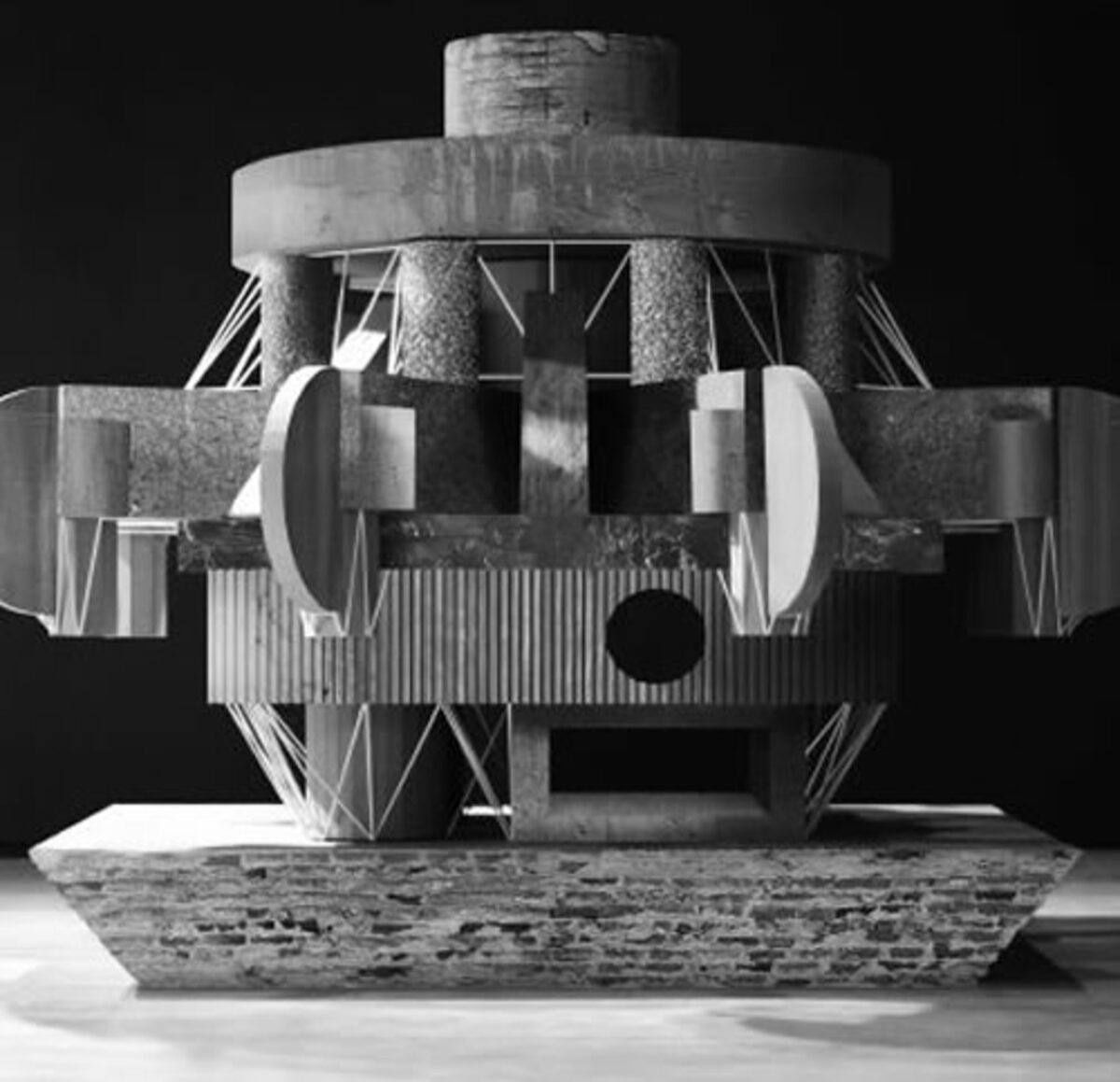 Black and white photograph of a geometric sculptural structure with cylindrical and angular forms, featuring stacked metal elements, curved discs, and triangular bracing against a dark background. The composition is symmetrical and frontal, showcasing industrial materials and architectural precision with strong contrasts of light and shadow.