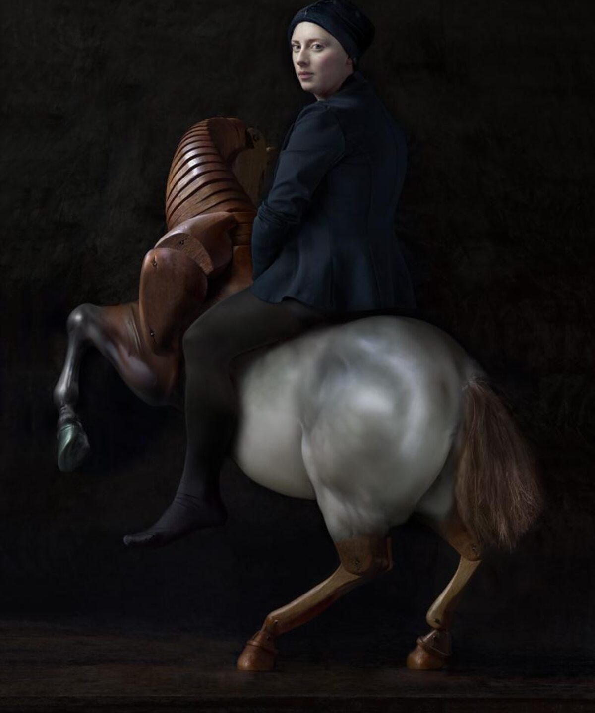 A woman in dark clothing and a dark headwrap sits atop a rearing wooden rocking horse against a black background. The horse, rendered in rich brown tones with a white blaze on its chest, is captured mid-motion, while the woman gazes toward the viewer with a composed expression.