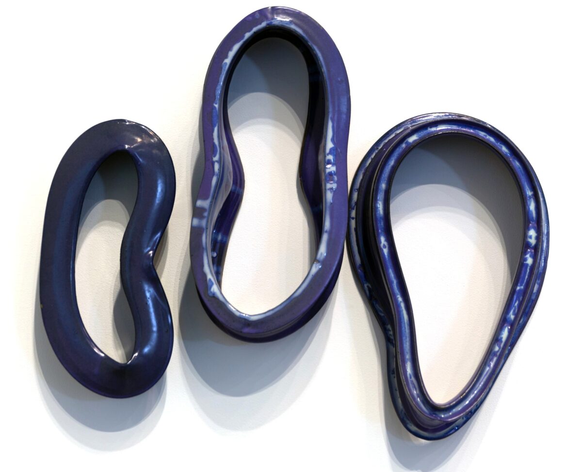 Three deep blue ceramic rings of varying sizes arranged in a row against a white wall, each casting a soft shadow. The glossy, glazed surfaces reflect light, revealing the organic, hand-formed quality of the sculptural objects.