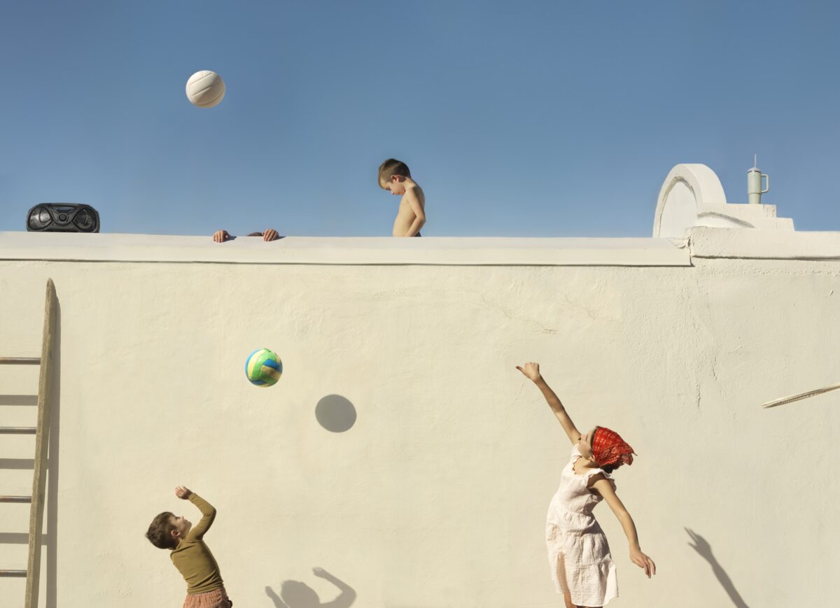 Children play with balls against a white concrete wall, with one child perched on top and others positioned below. The composition is divided between the bright blue sky above and the stark white architectural surface, creating a striking contrast that emphasizes the playful figures and their shadows cast on the wall.
