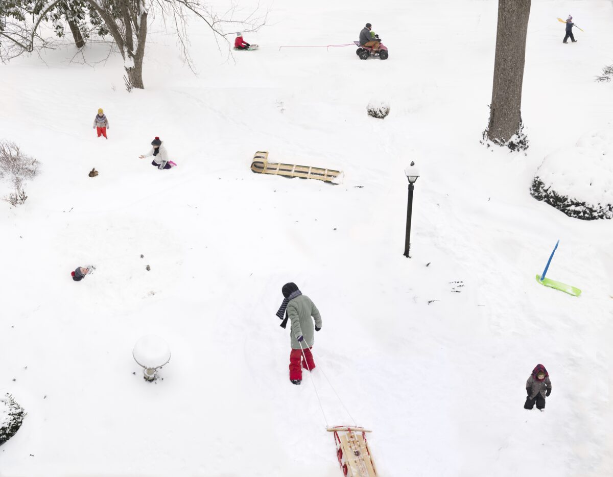 Aerial view of a snowy hillside with multiple people engaged in winter activities scattered across the landscape, including sledding, snowmobiling, and walking. The composition captures the scene from above, with bare trees, evergreen shrubs, and various wooden sleds distributed throughout the white expanse, creating a lively tableau of winter recreation.