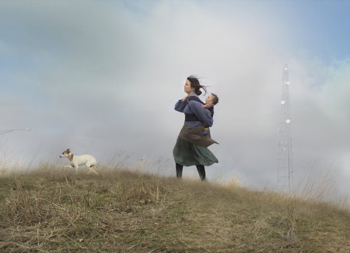 A woman holds a small child on a barren hilltop dotted with dry grass and vegetation, while a small dog stands nearby. A tall communication tower rises through the misty background on the right side of the composition, creating a stark contrast between the intimate human figures in the foreground and the industrial structure beyond.