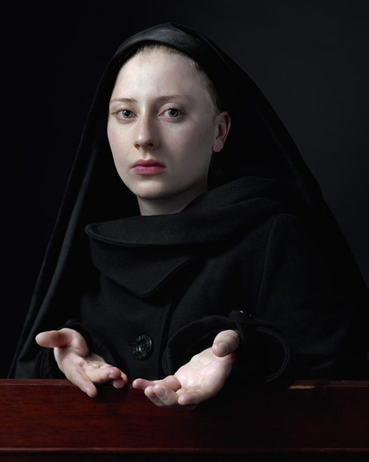 A pale-skinned figure wearing a black hooded cloak gazes directly forward with a neutral expression against a dark background. Their hands are positioned open and gesturing toward the viewer across a rich wooden surface, creating an intimate yet enigmatic portrait composition.