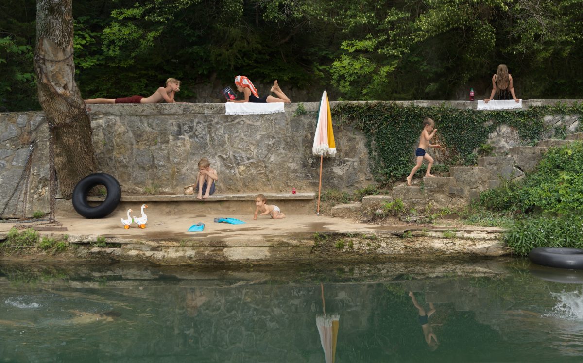 A leisurely riverside scene where multiple children and adults occupy a tiered stone embankment in various states of summer play and relaxation. Some figures recline on the upper ledge, others wade or sit along the sandy beach below, while a pair of white geese, beach toys, and an umbrella populate the foreground. Dense green foliage frames the scene, and the still water below mirrors the activity above.
