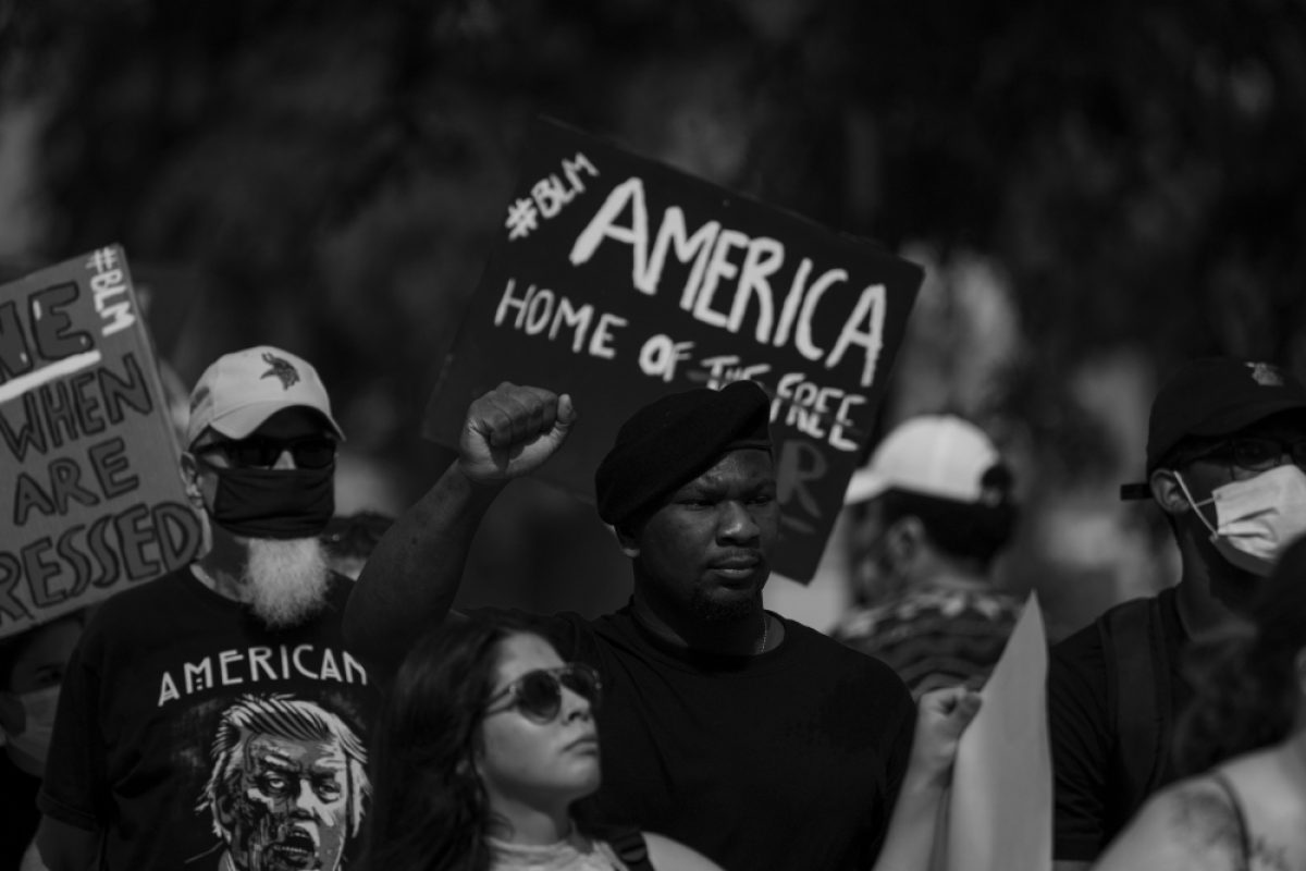 Black and white photograph of a protest crowd holding signs, including one reading "AMERICA HOME OF THE FREE." Participants wear masks and caps, with raised fists visible among the group, capturing a moment of civil demonstration and activism.