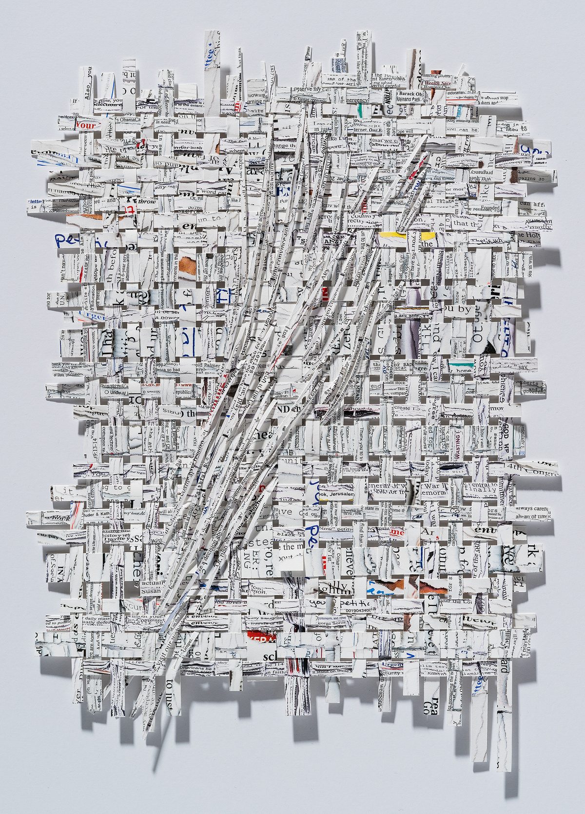 A three-dimensional collage of shredded and layered printed text and newspaper fragments arranged on a white wall, creating a dense, textured composition with diagonal striations and scattered edges. Occasional pops of color—including yellow, orange, red, and blue accents—punctuate the predominantly white and gray tones.