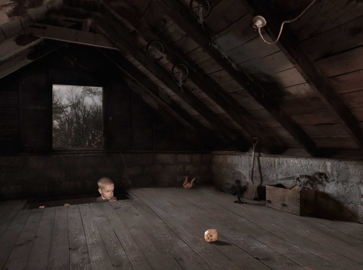 A young child peers over wooden floorboards in a sparse, dimly lit attic space, with bare wooden beams overhead and a window showing bare winter trees. A doll's head rests on the floor in the foreground, while another doll and various objects are scattered throughout the shadowy interior.