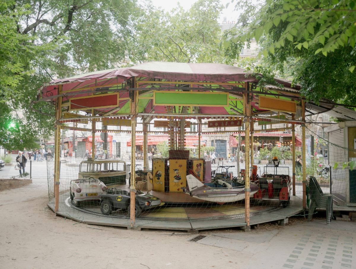 A weathered carousel with a faded red and yellow canopy sits in an urban plaza, its small vehicles painted with comic book characters including Batman. Surrounding trees frame the scene, with brick buildings and pedestrians visible in the soft, overcast light beyond.