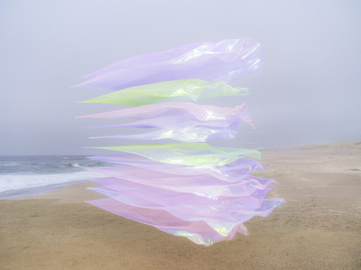 Translucent iridescent fabric strips in shades of lavender, pale green, and pink are layered and suspended vertically above sand, with an ocean and shoreline visible in the soft-focused background.