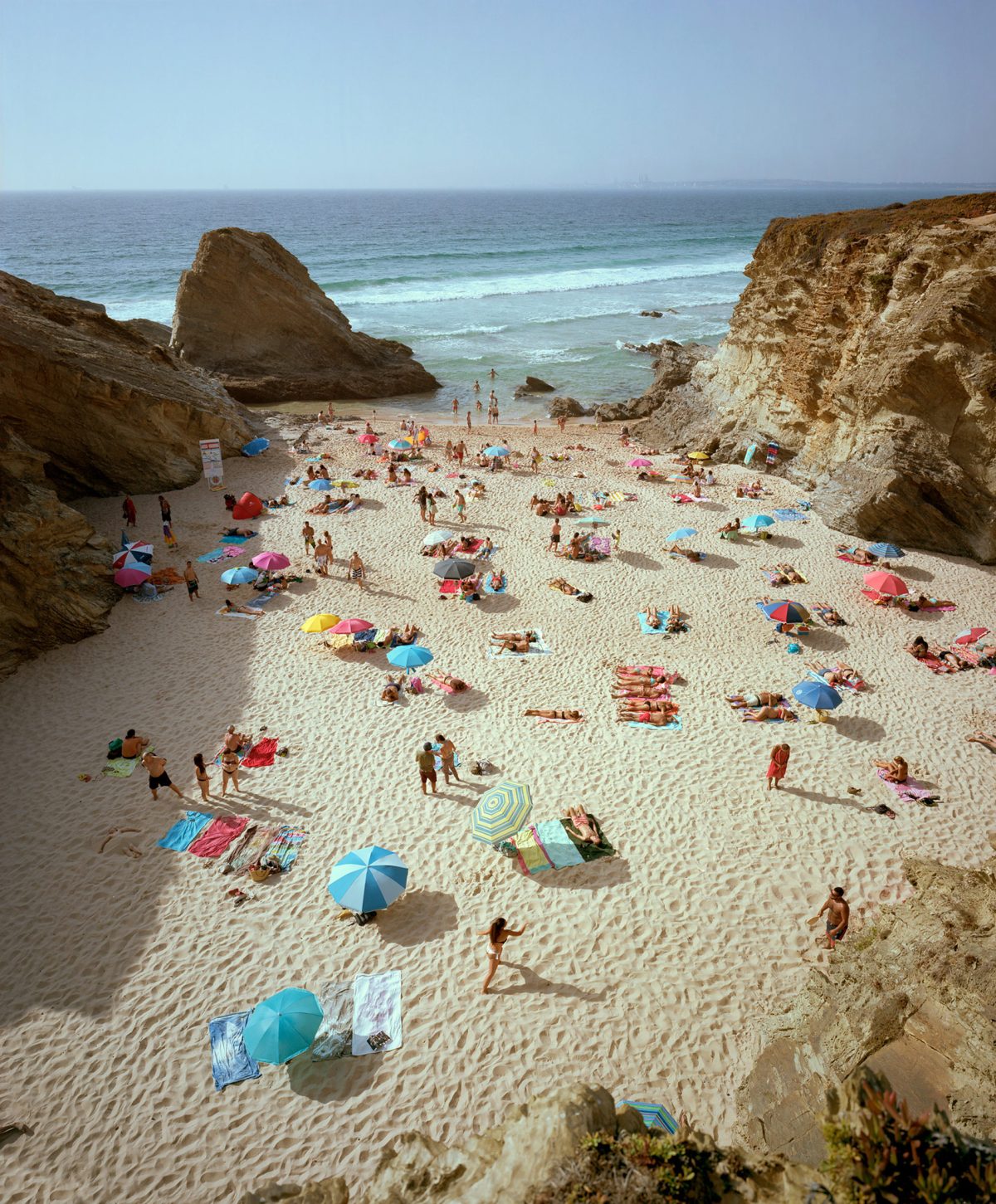 Aerial view of a crowded beach nestled between towering ochre-colored cliff formations, with numerous beachgoers scattered across pale sand beneath colorful umbrellas in shades of blue, pink, and yellow. Turquoise waves and rocky outcroppings frame the scene, while the vast horizon extends across a clear sky.