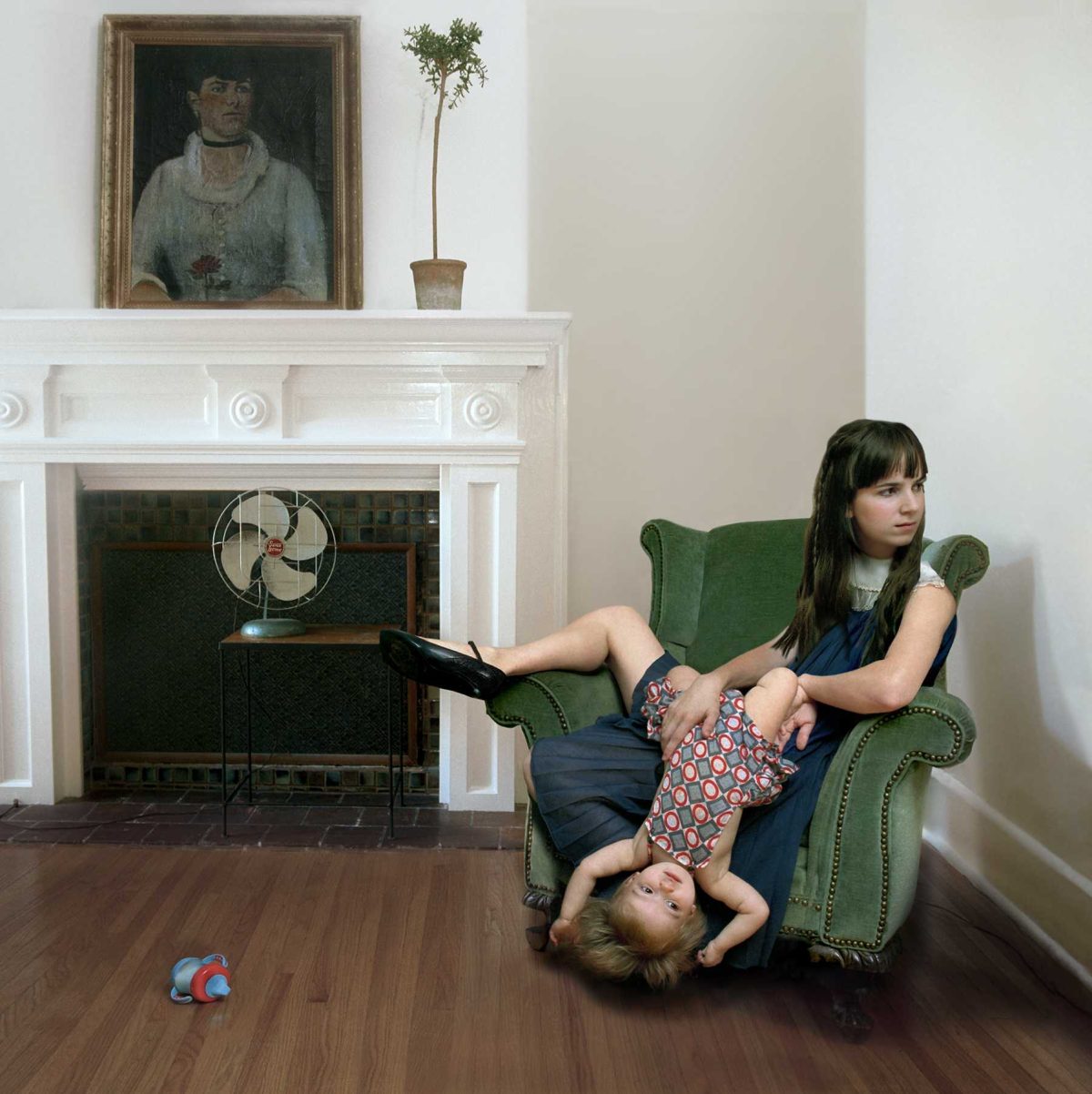A woman and young child sit together on a green upholstered armchair in a bright, classically decorated interior. The woman holds a book while the child reclines across her lap with legs extended. Behind them, a white fireplace mantel displays a framed portrait and a potted plant, with a vintage fan visible in the fireplace opening. Warm wood flooring and neutral walls frame the domestic scene.