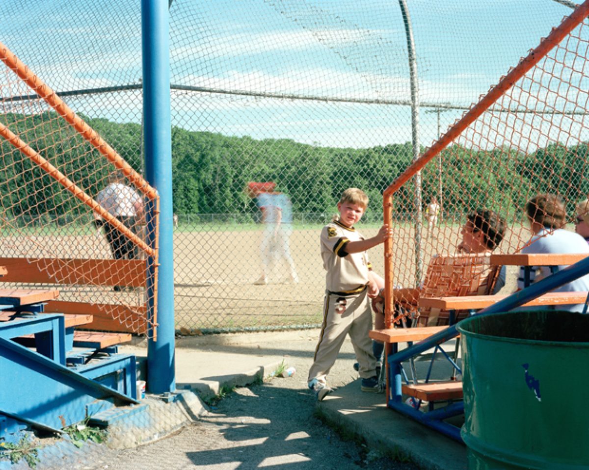 A baseball dugout scene viewed through a chain-link fence, with players and spectators in a sunny outdoor stadium. Vibrant primary colors—bright blue structural elements, orange and red netting, and green equipment—contrast against the natural tan infield and tree-lined background.